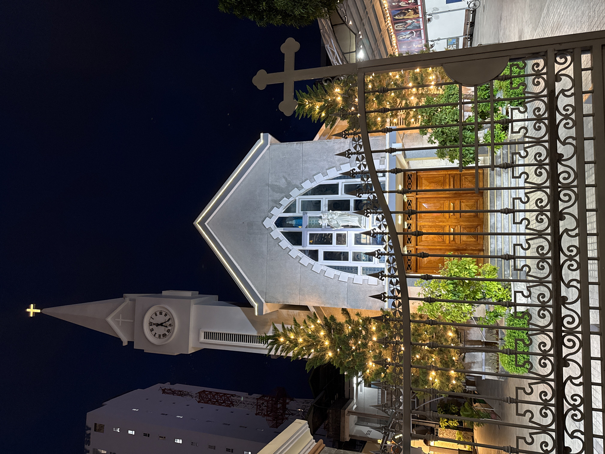 The image shows the front facade of a church at night. The church has a tall bell tower with a clock and a cross at the very top, illuminated by a yellow light. The church building is made of light-colored stone and has a distinctive arched window with stained glass. Two Christmas trees adorned with fairy lights are visible on either side of the entrance. A decorative metal gate with intricate scrollwork stands in front of the church, partially obscuring the view of the entrance, which features a double wooden door. A large, grey, standing cross is visible to the right of the church, behind the gate. A tall, modern building with illuminated windows and a red construction crane are visible to the left of the church. The sky is dark, indicating it is nighttime. On the right side of the image, beyond the church, there appears to be an outdoor area with a covered structure and a series of religious paintings or icons displayed on a wall. A bicycle is parked in the distance. The scene is likely festive, given the Christmas trees and lights.
The church is identified as the Nha Trang Cathedral, also known as the Christ the King Cathedral, located in the city of Nha Trang, Vietnam. It is a Roman Catholic cathedral built in a Gothic architectural style.