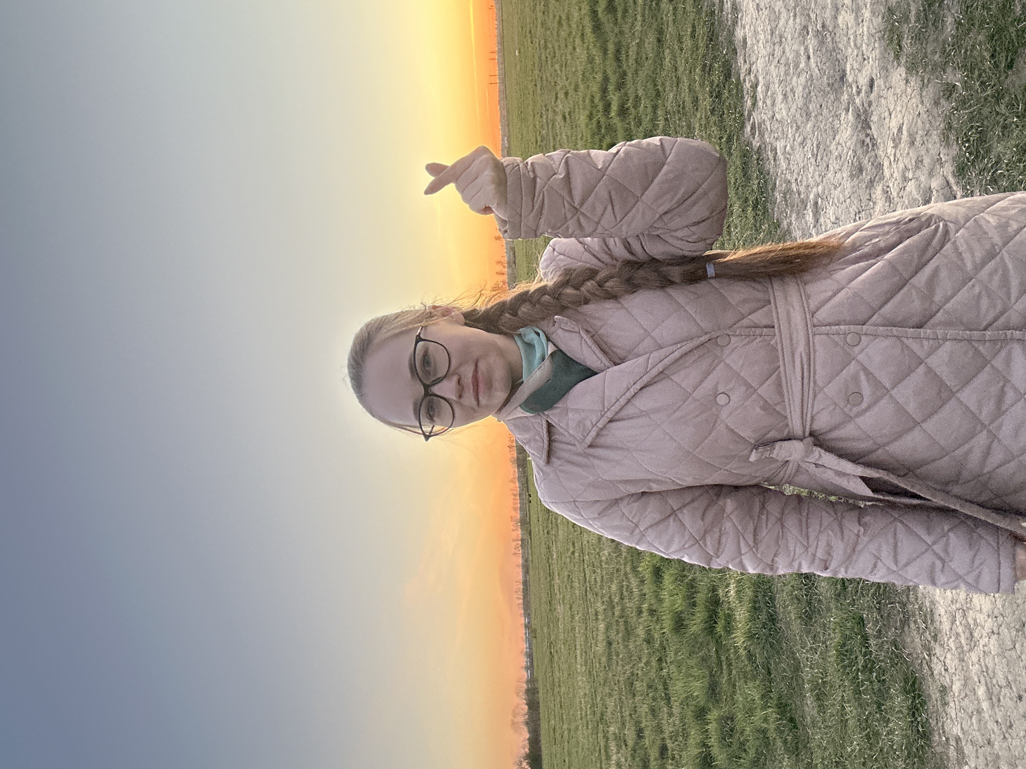 A young woman with blonde hair braided over her right shoulder stands outdoors, making a finger heart gesture towards the sunset. She is wearing glasses and a light pink quilted coat with a tie belt. The background shows a field of green grass. The sky is a gradient of orange to blue, indicating sunrise or sunset. The location appears to be a parkland. Due to the lack of contextual clues, it is difficult to confirm this picture was taken in Saint Petersburg, Russia.