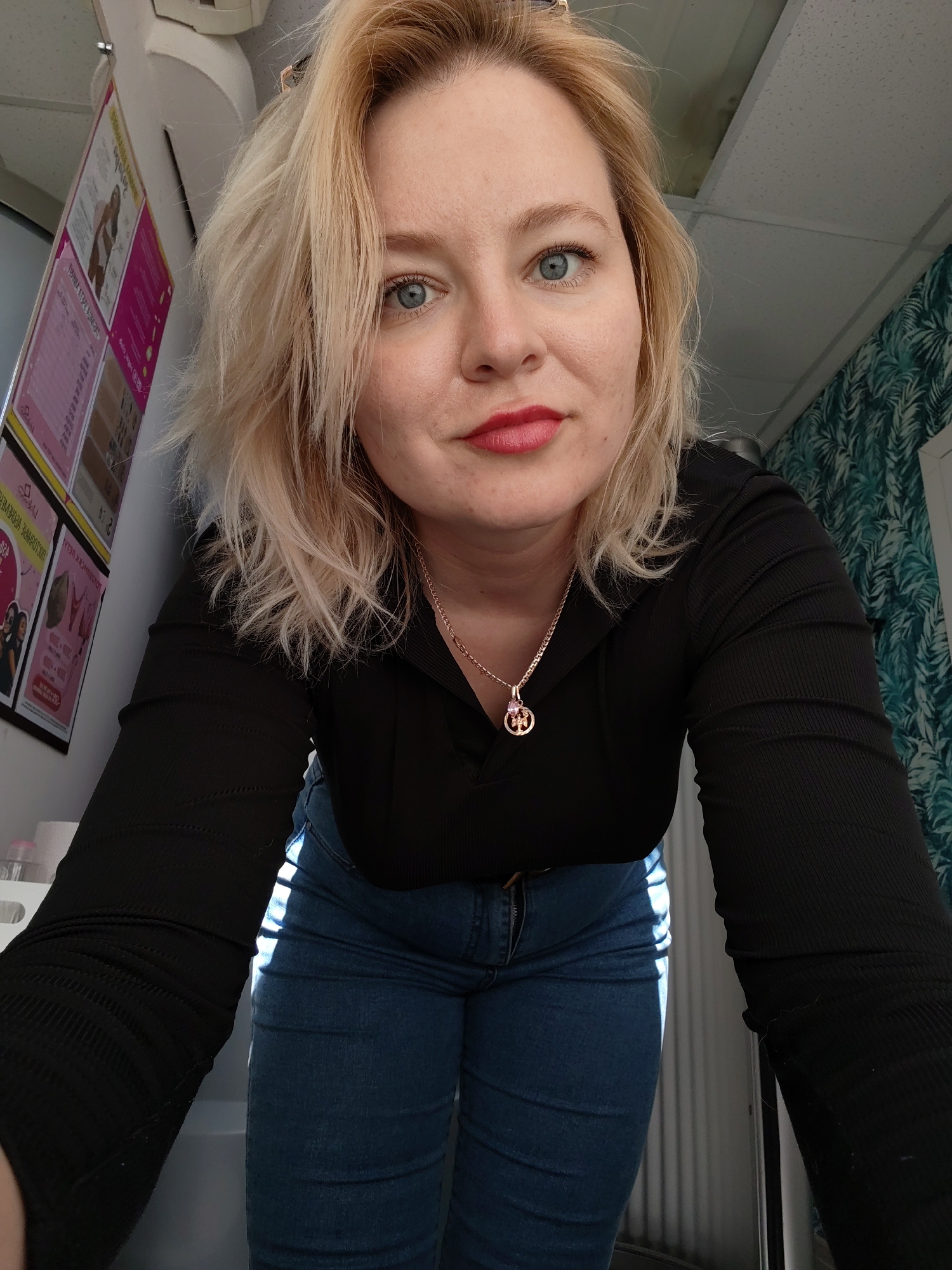 A Caucasian woman with blonde hair and blue eyes is facing the camera. She is wearing a black long-sleeved shirt and blue jeans, with a gold necklace. She is indoors. Behind her on the left is a framed poster with images and text, partially obscured. To the right is a wall covered in a dark green and blue tropical-themed wallpaper. The location is in Izhevsk, Russia.