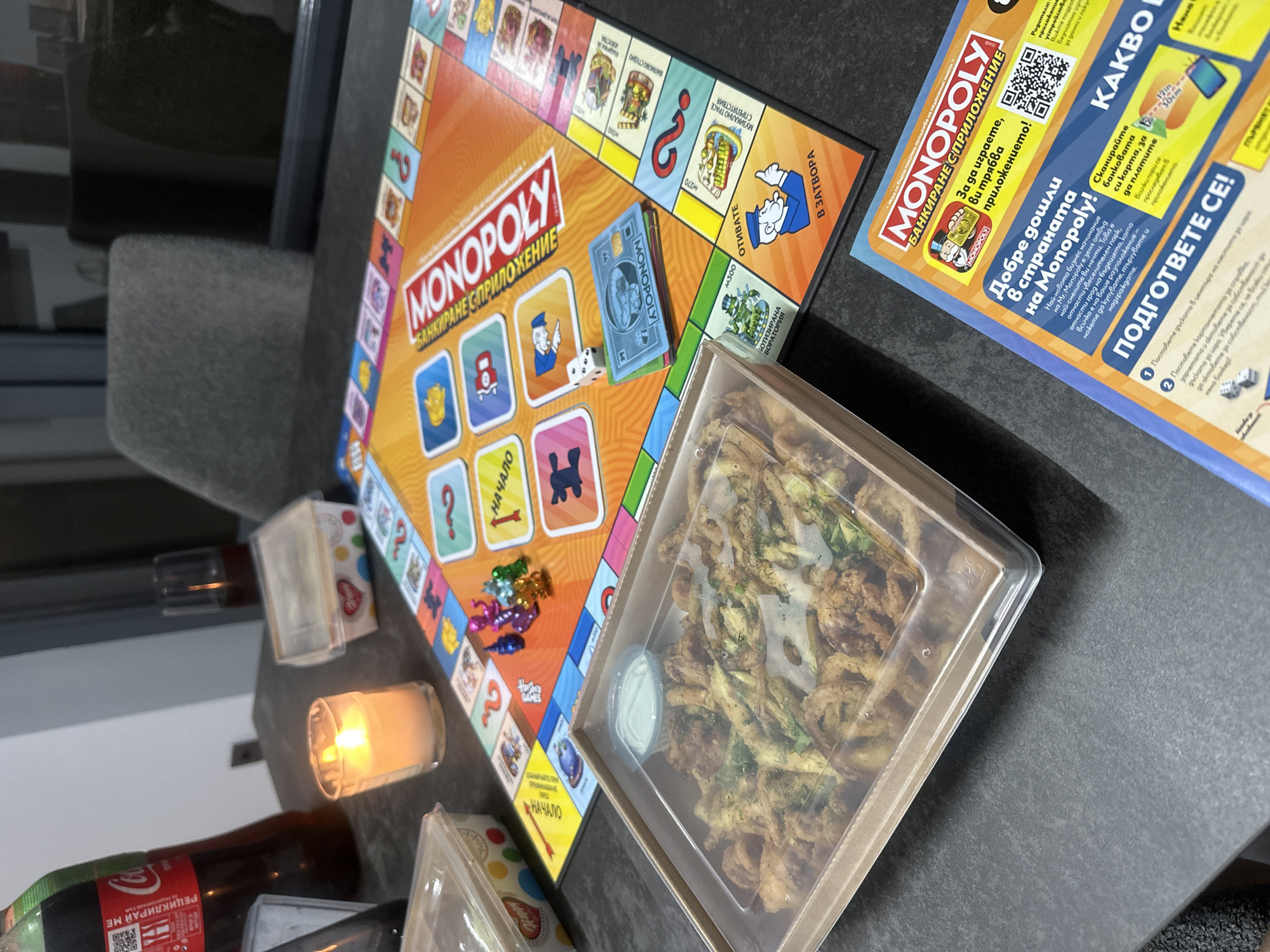 The image shows a close-up, eye-level view of a Monopoly board game spread out on a dark, textured surface. To the left, a takeout container filled with fried food and a small cup of dipping sauce is placed in the foreground. A lit candle in a glass holder sits behind the food container, casting a warm glow. A bottle of Coca-Cola is visible in the upper left corner of the frame, and a glass of dark liquid is to the right of the candle. The Monopoly board itself is partially visible, featuring colorful spaces, game tokens (including small dinosaur figures), dice, and what appears to be Monopoly money or cards. The text on the board is in Bulgarian. The background is blurred, showing a glimpse of a dark chair and reflective surfaces, suggesting an indoor setting. The overall scene suggests a casual game night with food and drinks.