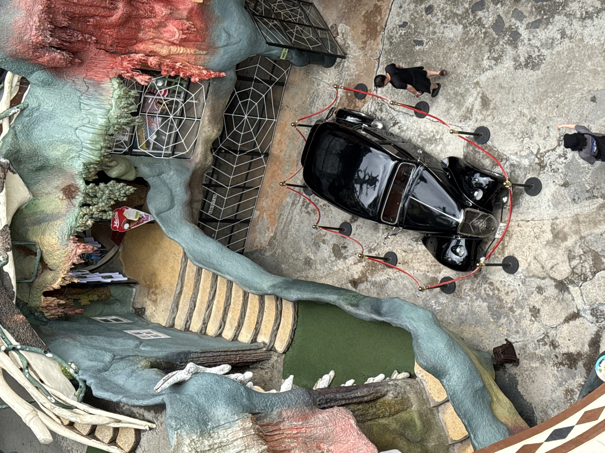 The image is an overhead view of a display at the Crazy House in Da Lat, Vietnam.
The display features a miniature, fantasy-like environment, with elements of organic shapes, artificial textures, and structural components. A black vintage car is centrally positioned on a concrete-like surface, enclosed by red stanchion ropes and black posts. Two individuals are visible near the car, one in a black outfit and the other in gray, appearing to be interacting with the display or each other.
The environment includes a section with building facades, a set of stairs leading to an area with artificial green grass, and a section of white, bone-like structures. The backdrop consists of a mix of rough, colorful surfaces with textures mimicking natural formations. There are some grid-like structures adjacent to the car that create the impression of a building exterior. The surrounding ground appears to be a concrete surface with variations in color and texture.