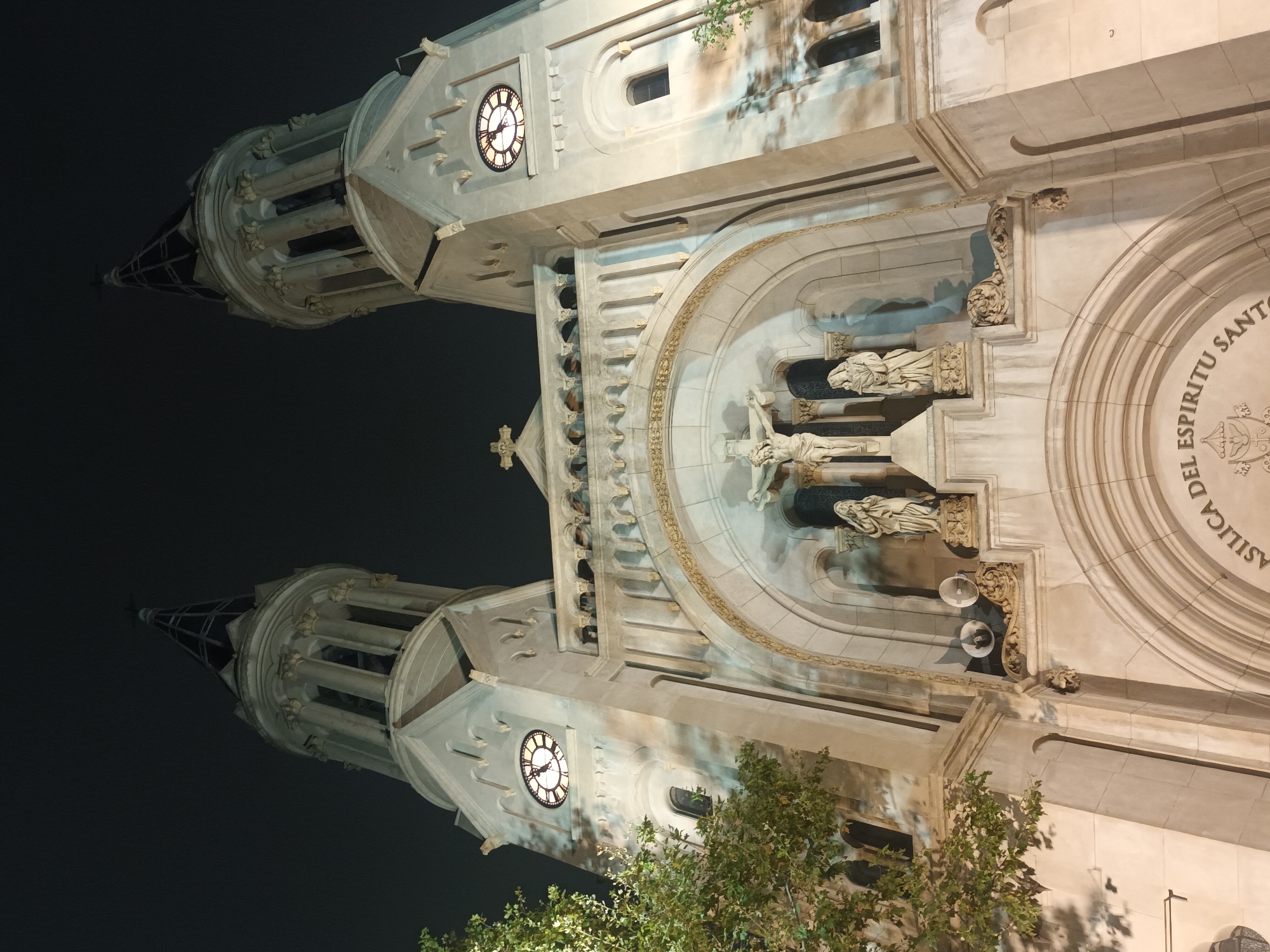 A low-angle, rotated, nighttime shot of the Basilica del Espiritu Santo, located in the Belgrano neighborhood of Buenos Aires, Argentina. The facade of the building is beige and includes two bell towers with clocks. A statue of Jesus on the cross is mounted above the main entrance. Statues of two other figures flank Jesus. A tree with green leaves is in the foreground. The night sky is dark.