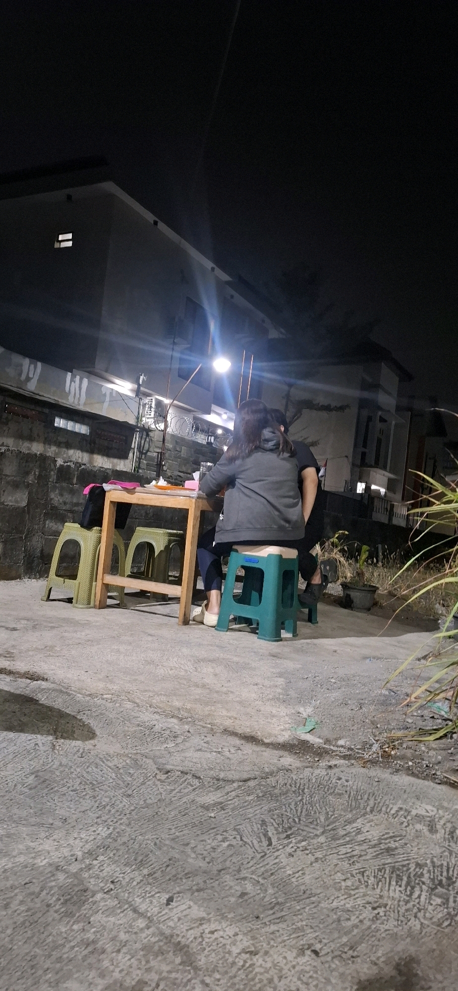 At night, two individuals are seated at a wooden table on a concrete outdoor patio, likely in Gajahan, Indonesia. One person, viewed from the back, is wearing a dark gray hooded jacket and dark pants, and is seated on a teal plastic stool. The other person, also seen from the back, is wearing a dark shirt and is seated on a dark stool. A smaller wooden table is positioned near the main table, and there are two light green plastic stools adjacent to it. On the table, various items are visible, including plates and glasses. The background features a grey stone wall, a building with lights, and some greenery. The overall lighting suggests an outdoor dining or gathering space at night.