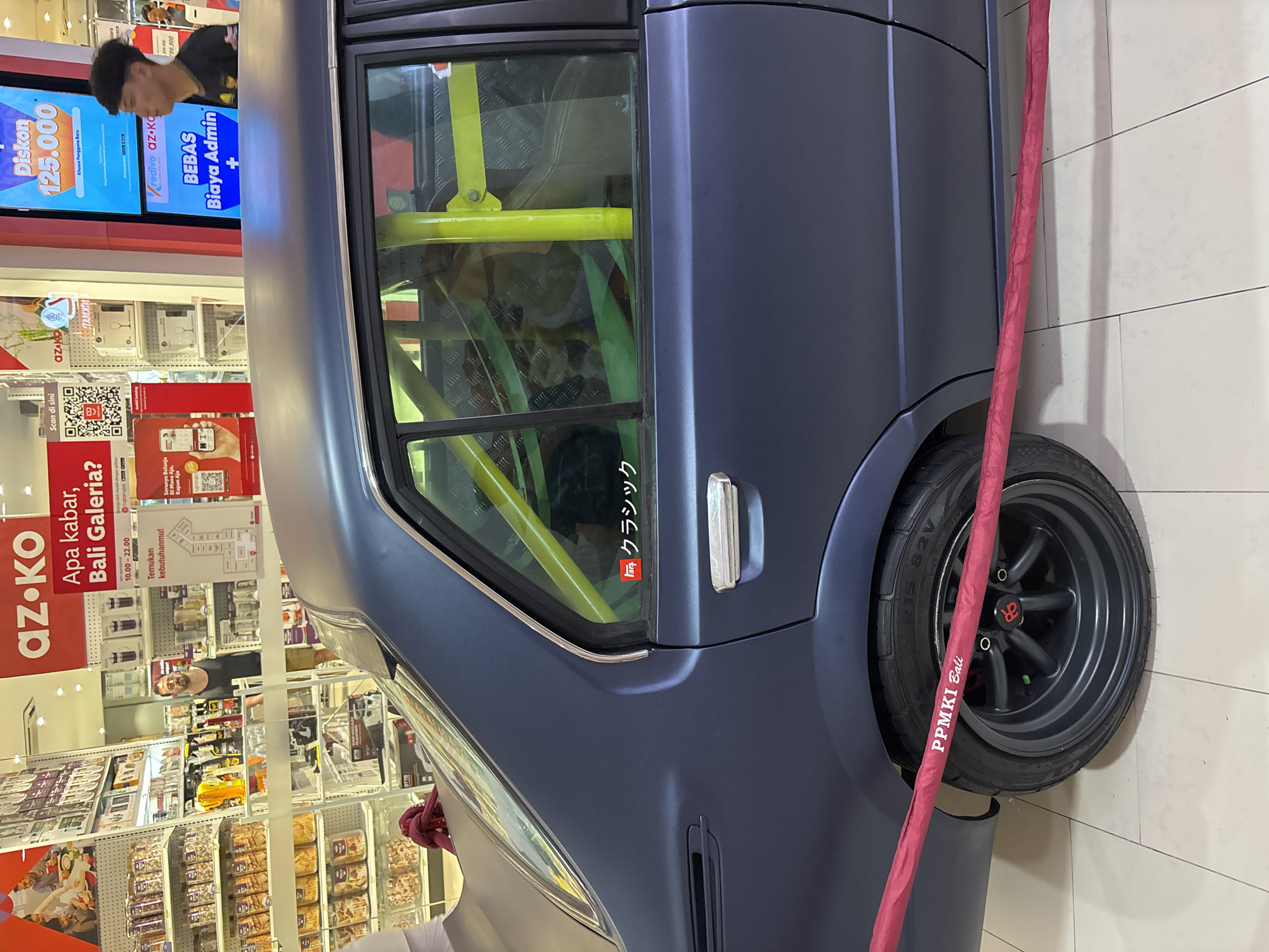 The image shows the side of a matte blue car parked indoors, likely in a shopping mall. The car has a yellow roll cage visible through the window, with a Japanese character sticker on the door. The rear wheel is black with a red center cap, and a red umbrella leans against the tire. A shopping center is visible in the background with various displays and shoppers. The mall appears to be in Bali Galeria, Kuta, Indonesia, based on the "az.ko Bali Galeria" signage and "PPMKI Bali" on the umbrella. There are promotional displays showing discounts.
