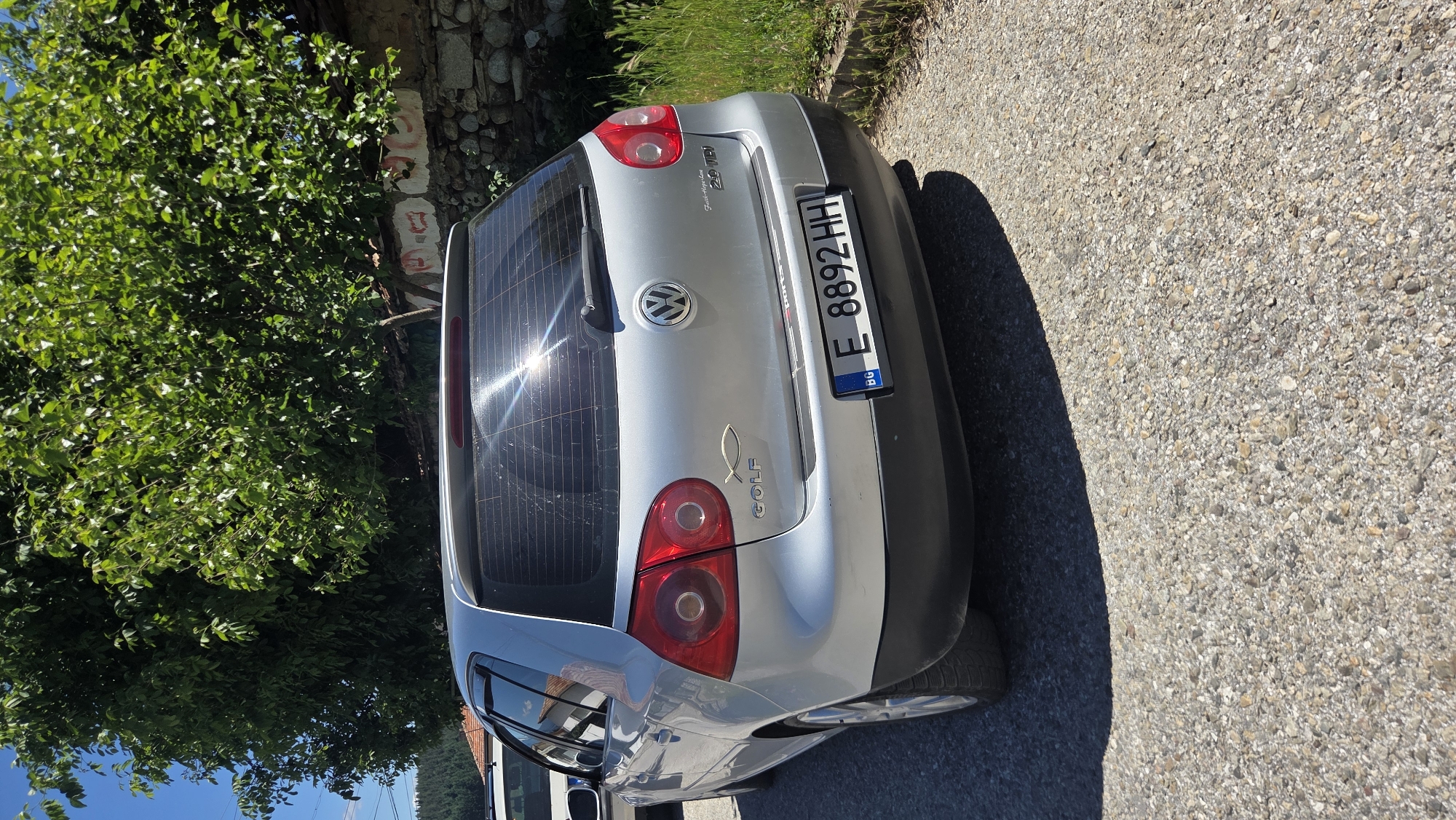 A silver Volkswagen Golf is parked on a gravel road. The car is viewed from the rear passenger side. It has a license plate that reads "E 8892 HH" and a "BG" sticker on the bottom left. A fish symbol is on the left side of the trunk, below the "Golf" text. The car is parked in front of a stone wall covered with some graffiti. Lush green trees and grass surround the area, with a clear blue sky above. The sun is shining brightly, creating a glare on the car's rear window.