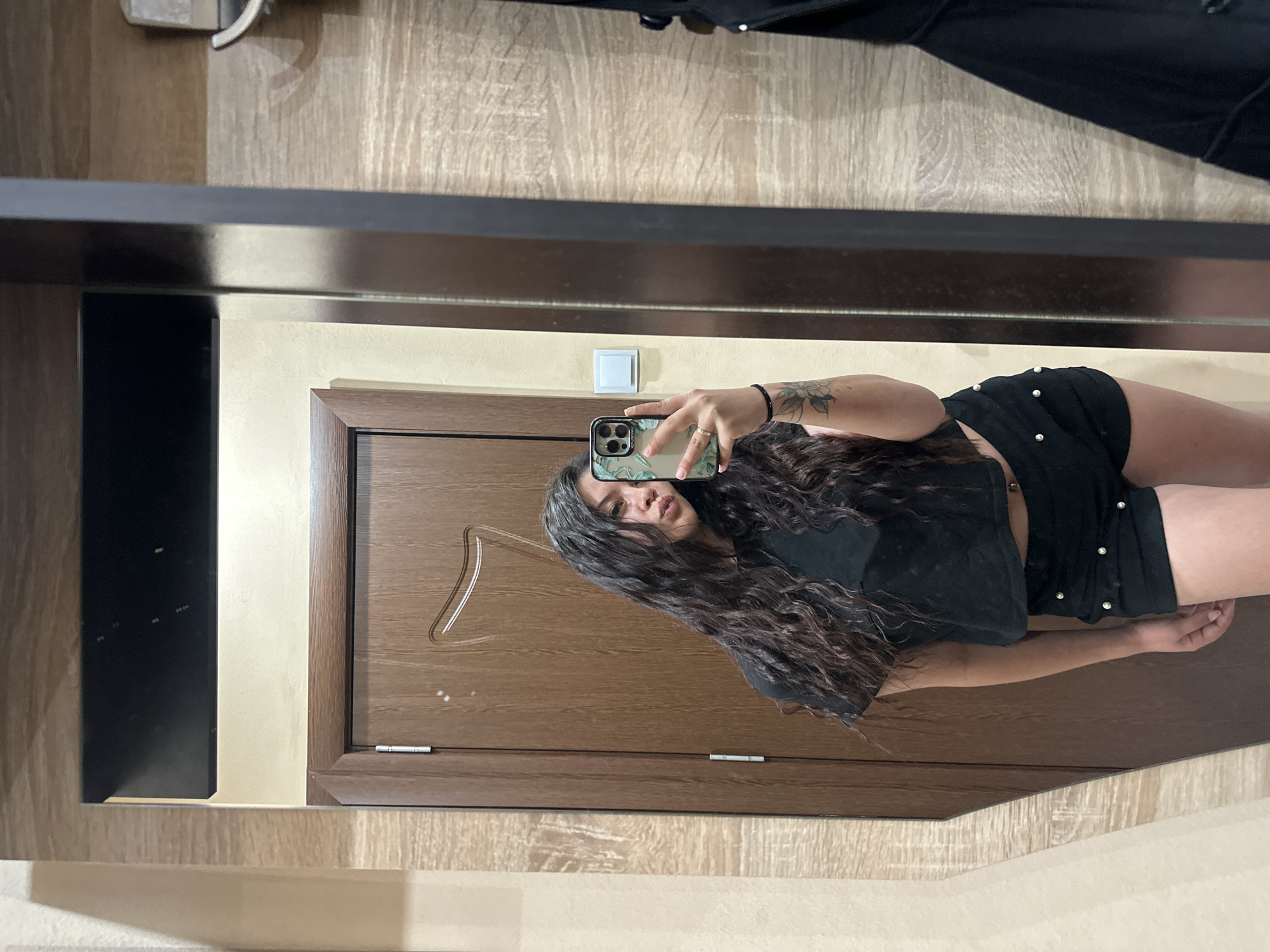 A young woman with long, dark, curly hair is taking a mirror selfie in a room. She is wearing a black crop top and black shorts adorned with pearl-like studs. She has a tattoo on her left arm and is holding a smartphone with a floral case. The background features a wooden door, a beige wall, and what appears to be a closet or wardrobe with a black garment hanging on it. The lighting suggests an indoor setting. The image does not contain information that would allow for precise identification of the city of Sliven, Bulgaria.