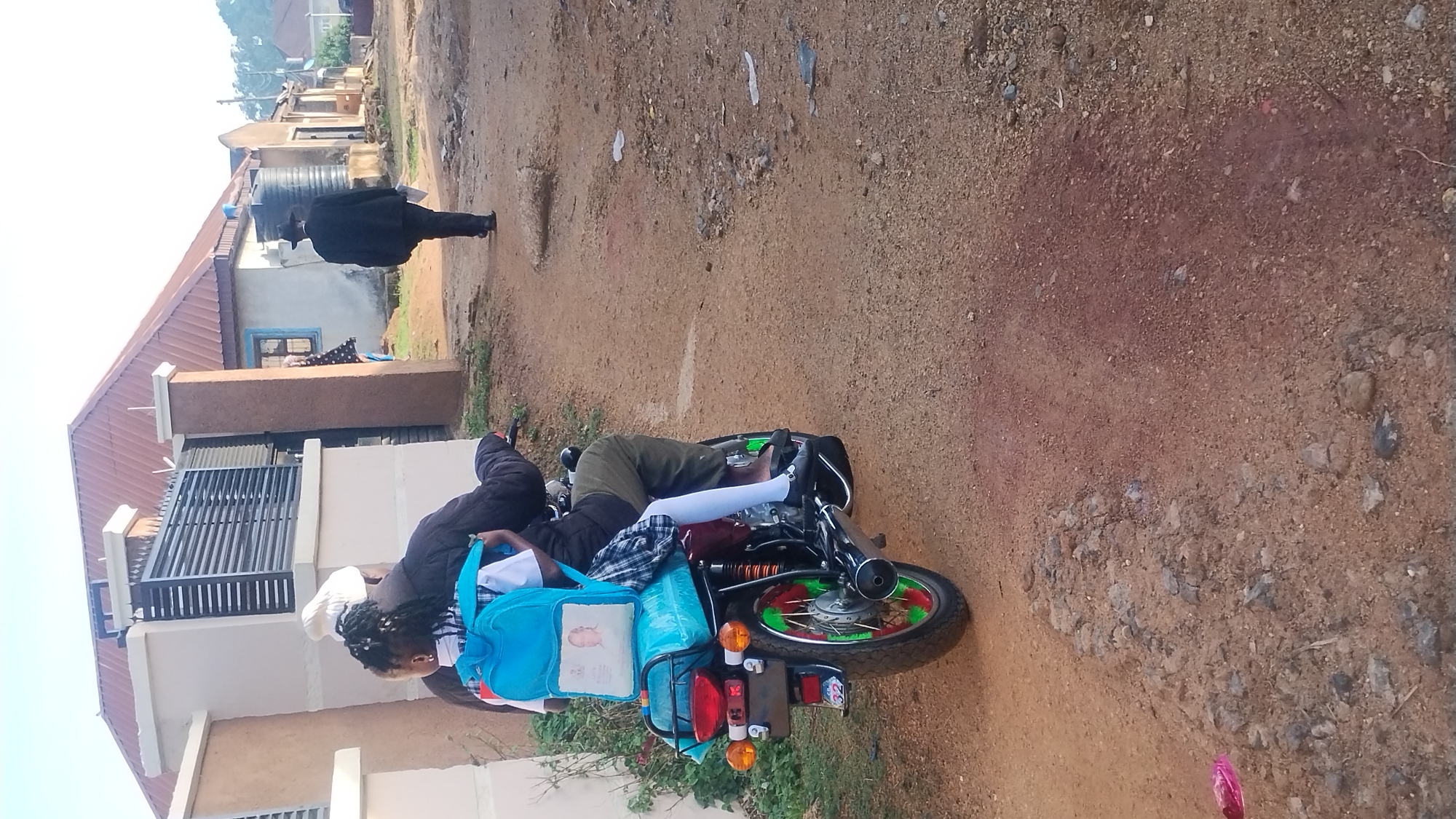 The image depicts a daytime scene on an unpaved, reddish-brown dirt road in what appears to be a residential area. The sky is bright and clear.
In the foreground, two individuals are positioned near a motorcycle. One person is seated on the motorcycle, wearing dark pants, a dark jacket, and white socks. Their feet are on the footpegs. Another person, likely a child with braided hair, stands next to the motorcycle, facing away from the viewer. This child is carrying a light blue backpack with a light-colored patch featuring an unidentifiable image and some faint, illegible text. A similar blue bag is secured to the back seat of the motorcycle. The motorcycle itself has distinctive bright green and red accents on its front wheel spokes. The number "32" is visible on its rear license plate.
In the mid-ground, a person wearing a dark jacket and a dark hat walks along the dirt road, heading away from the foreground subjects.
The background features several single-story residential buildings with corrugated iron roofs. One building has a large black water tank on its roof. The overall setting suggests a rural or semi-urban environment, possibly in a developing country. No specific emotions are discernible from the individuals.