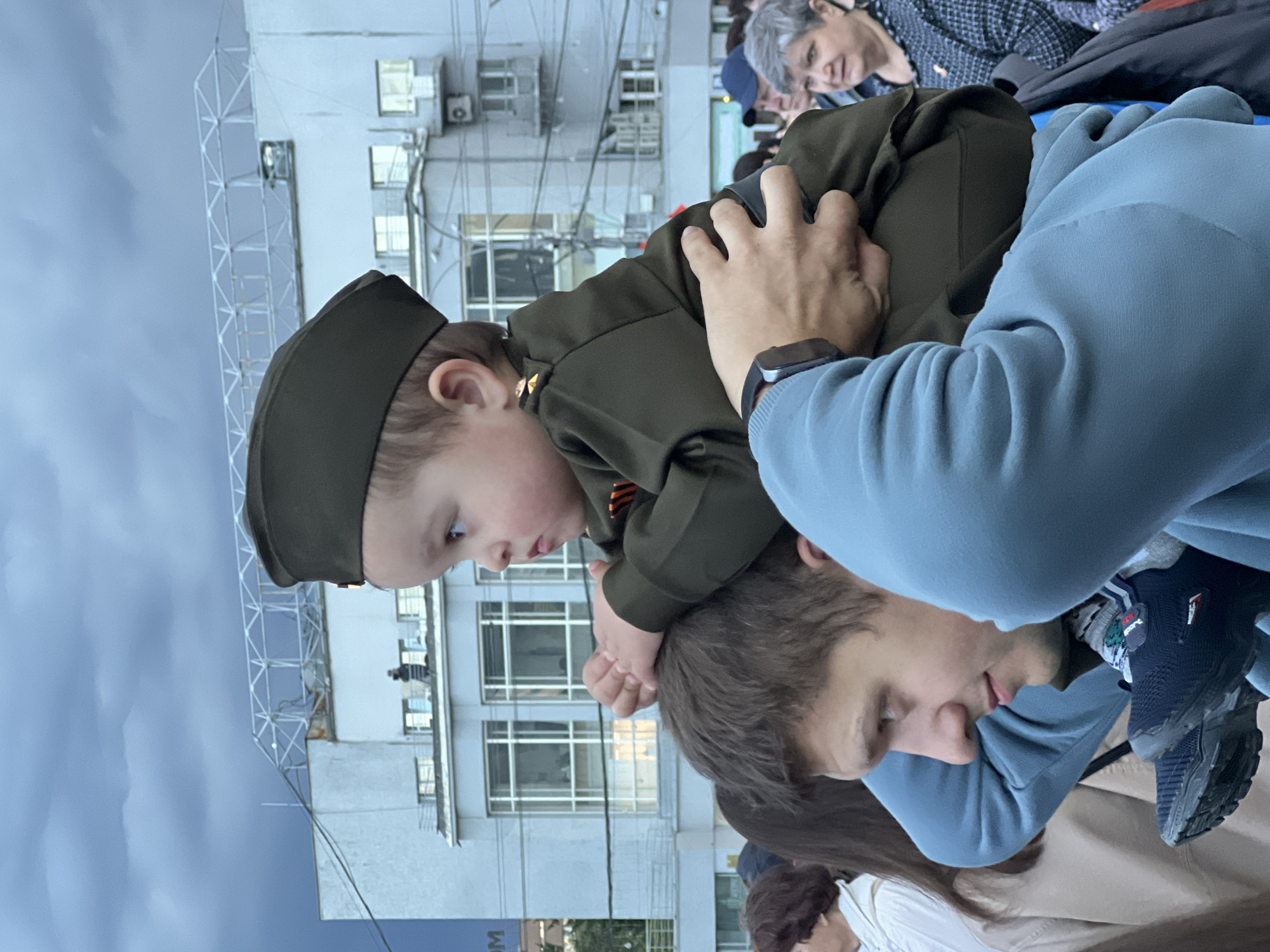 A young child, dressed in a military-style uniform and hat, is sitting on a man's shoulders outdoors. The man is wearing a light blue hooded sweatshirt. The child's hands are resting on the man's head, and the man's hand is resting on the child's back. The child is looking to the left. In the background, there is a multi-story building with numerous windows and air conditioning units, as well as a metal structure. The sky is overcast. There are other people visible in the surrounding area. This is likely a photo from the 2023 Victory Day parade in Novosibirsk, Russia.