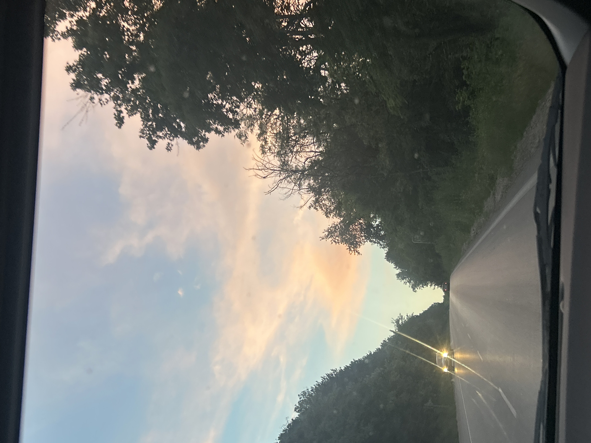 An image taken from inside a vehicle shows a road with trees on either side. The sky is a gradient of blue, pink, and orange, suggesting either sunrise or sunset. Two cars are visible on the road, one in the foreground with its headlights on, and another further down the road. The road appears to be a two-lane highway. The overall scene is serene and natural, with the sky and trees creating a picturesque backdrop.