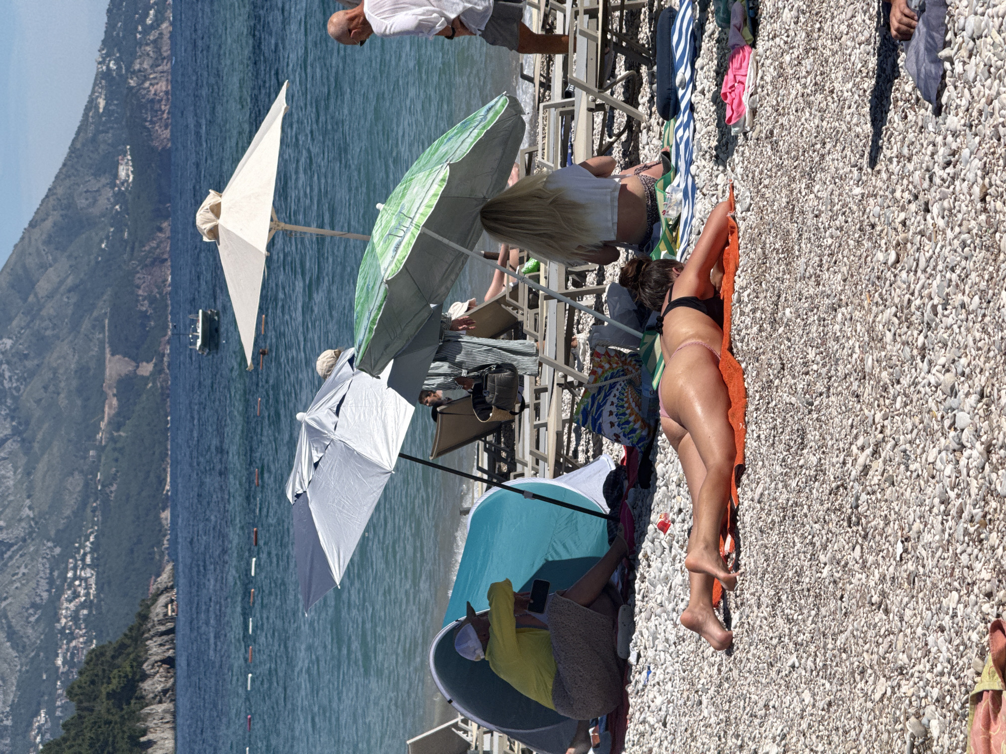 The image depicts a sunny day at a pebble beach in Budva, Montenegro, with the Adriatic Sea and mountains forming the backdrop. In the foreground, a person is sunbathing on an orange towel, wearing a black bikini. To the left, another person wearing a yellow shirt and a patterned skirt is seated under a light blue beach tent, holding a black smartphone. Further back, two individuals are seated under a large green umbrella, with one person displaying blonde hair and a white top. A man in a white shirt and grey shorts stands near the water's edge. Several beach chairs are visible, some unoccupied, and beach umbrellas offer shade. The sea is calm with a few boats in the distance. The overall scene portrays a typical beach day with people enjoying the sun and sea.