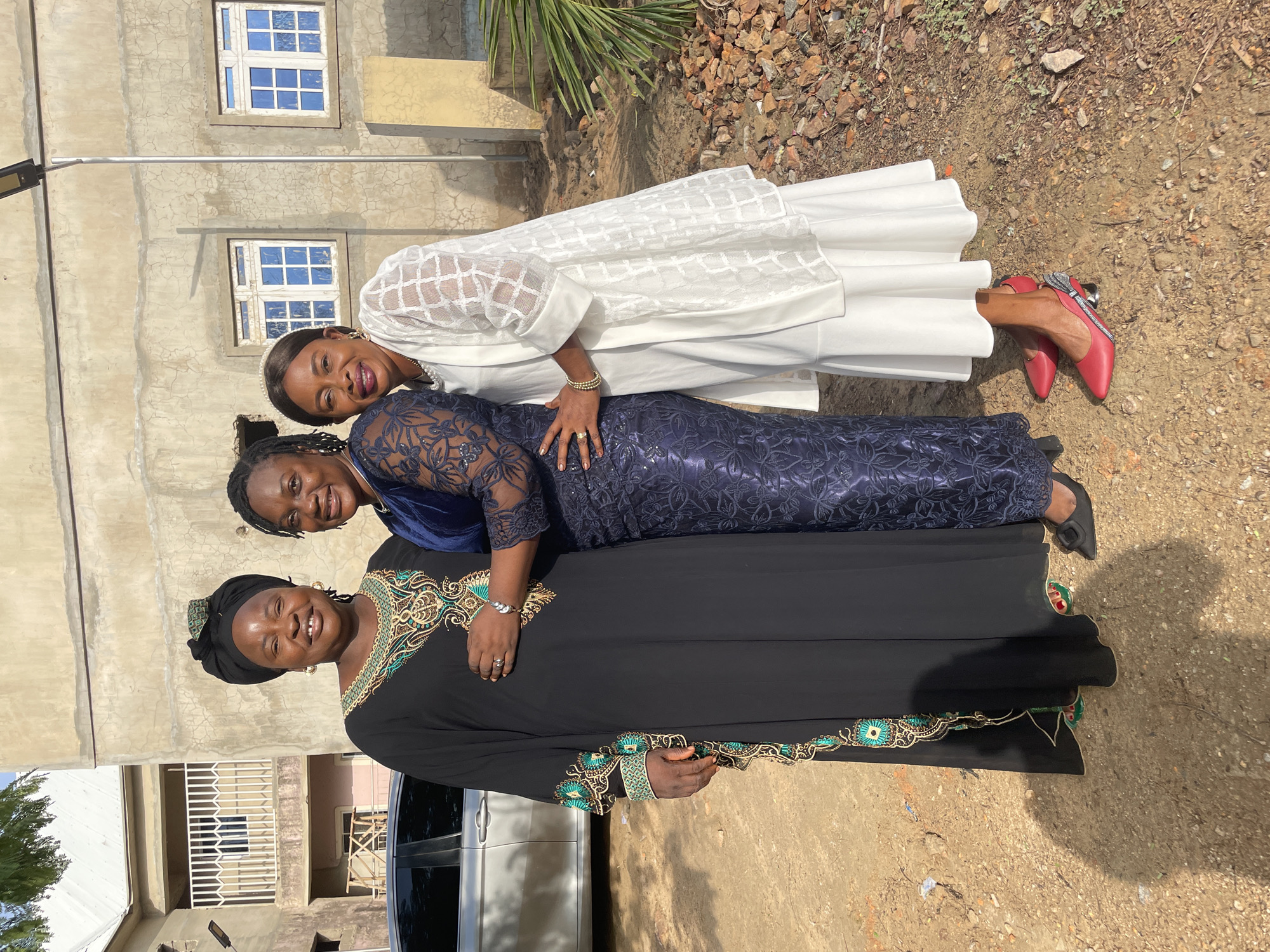 Three women are standing together, with the woman in the white dress leaning on the woman in the blue dress, who is leaning on the woman in the black dress. They are outside in Akwanga, Nigeria, with a building and car visible in the background. The ground is dirt. The women are dressed in elegant, formal gowns. The woman in the white dress is wearing red shoes.