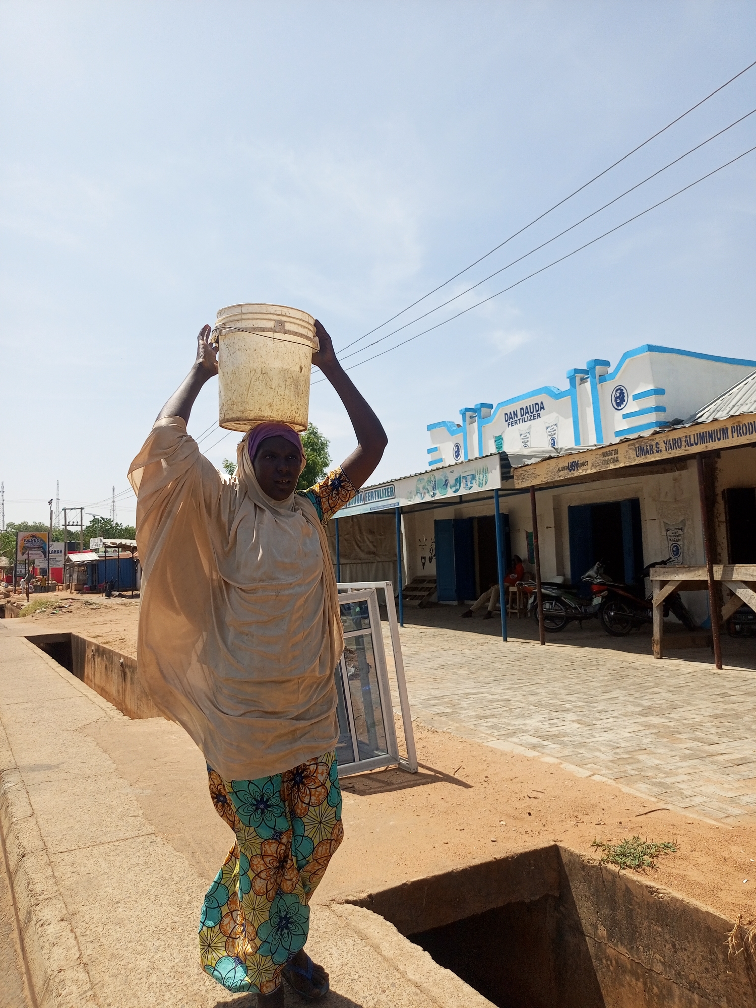 A woman carries a bucket on her head in an urban area. She is wearing a beige robe over a long-sleeved top with a colorful floral pattern and sandals. She is walking along a sidewalk with a drainage ditch. In the background is a building with a sign that reads "DAN DAUDA FERTILIZER" and "UMAR S. YARO ALUMINIUM PRODUCT." A person and a motorcycle are under the building's awning. Power lines stretch across the sky.