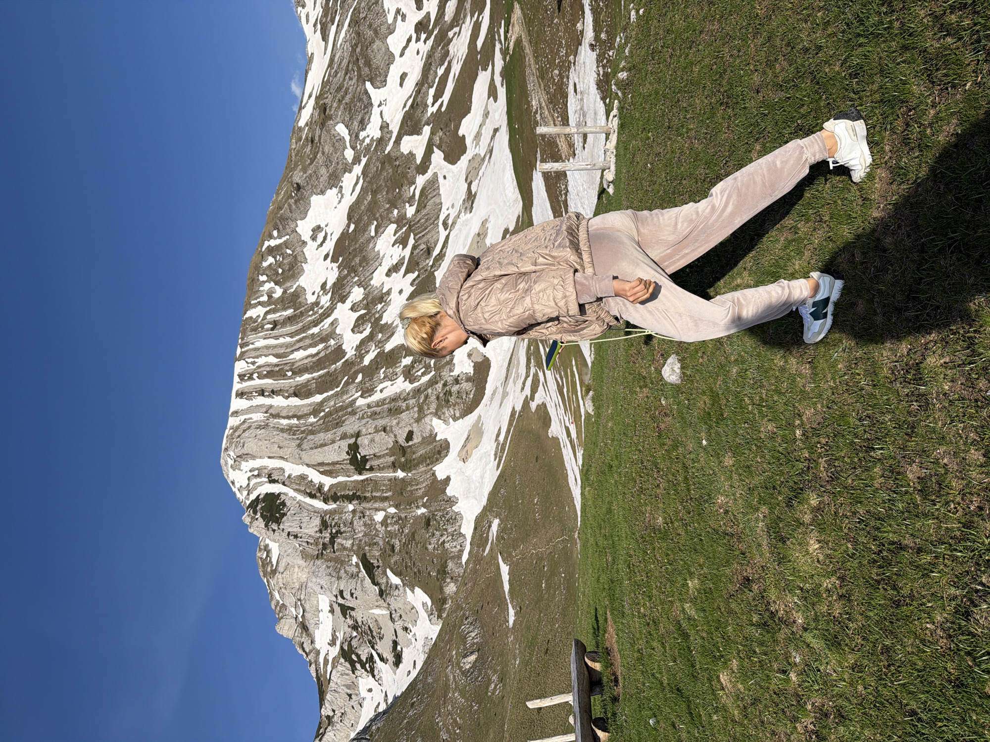 A woman lies on a grassy hillside in Боричје, Montenegro. She wears a quilted jacket and matching pants, with white sneakers on her feet. Behind her is a large mountain with patches of snow. The sky is clear and blue. A wooden bench is partially visible at the edge of the frame.