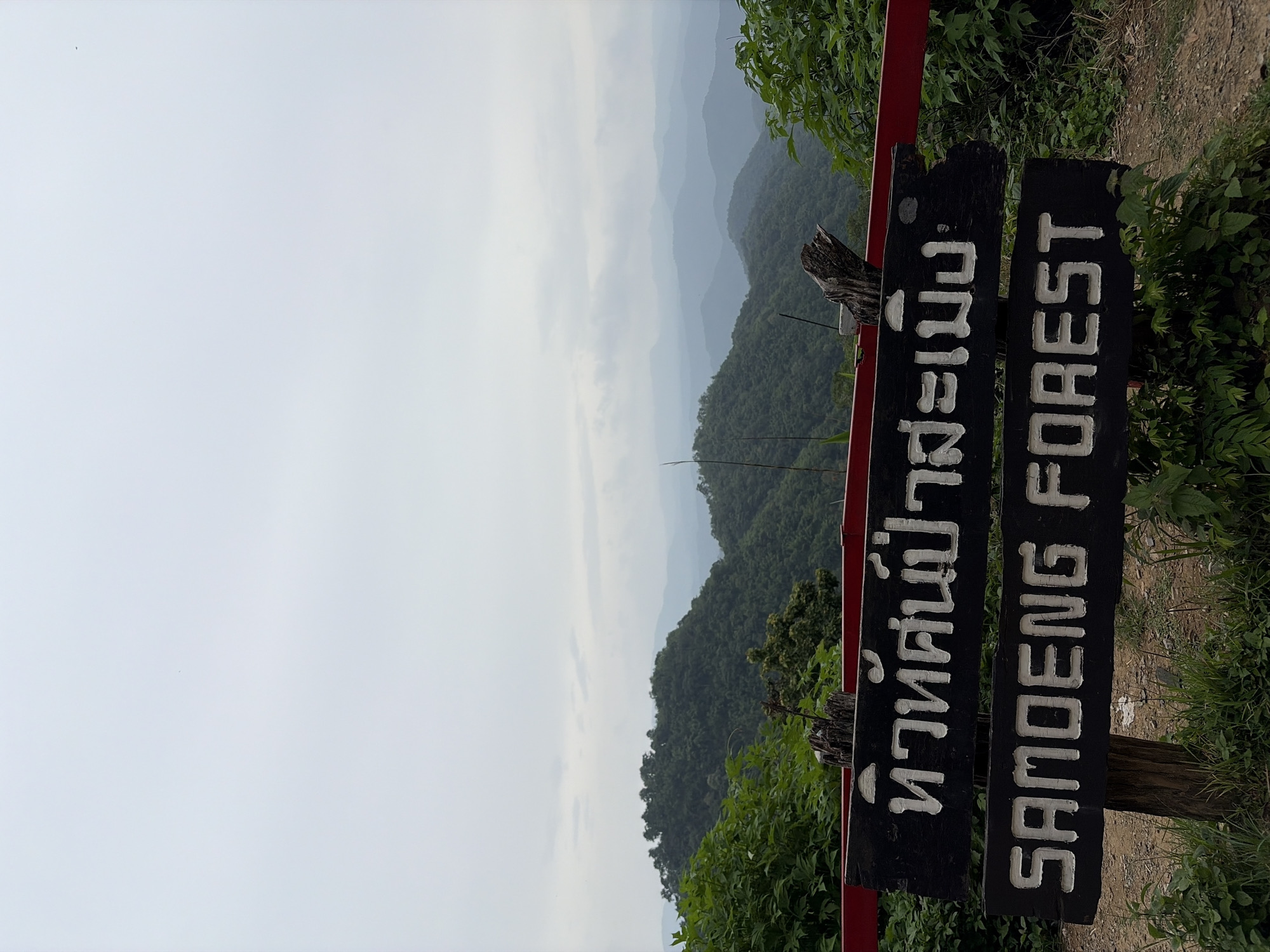 The image shows a wooden sign with white lettering in front of a misty mountain landscape. The top line of the sign contains Thai script, which reads "ห้วยศก์ป่าสะเมิง" (Huai Sok Pa Samoeng). The bottom line of the sign reads "SAMOENG FOREST" in English. The sign is mounted on two wooden posts. In the background, there are rolling green hills covered in lush vegetation, with several layers of mountains fading into the distance under a pale, overcast sky. To the left and right of the sign, there are green bushes and trees. A red railing is visible at the top of the sign. The location is identified as Samoeng Tai, Thailand.