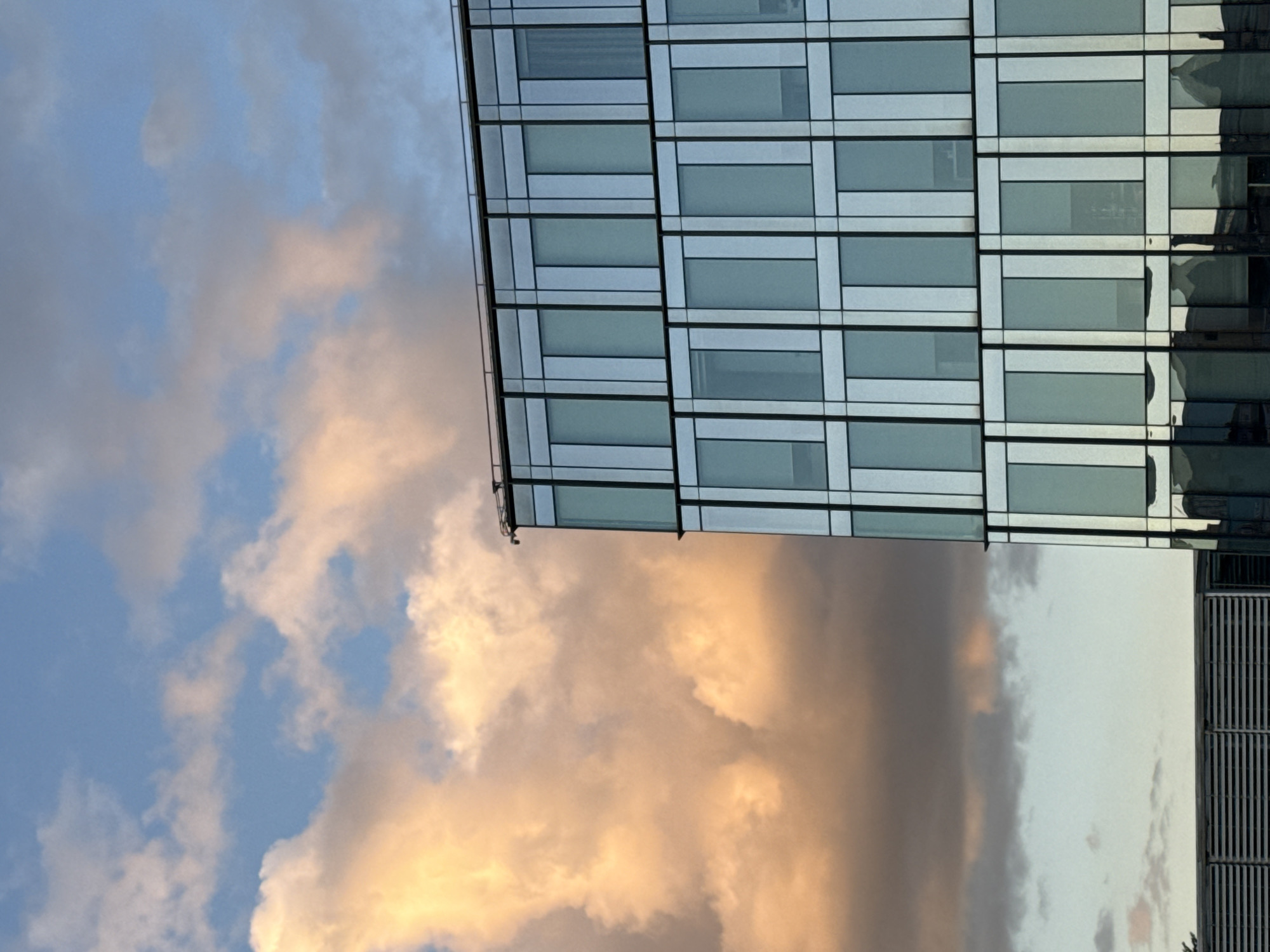 The image shows a modern glass-clad building in Moscow, Russia. The building has a rectangular grid pattern of windows. The sky is filled with clouds, some illuminated by the setting sun in golden hues, contrasting with areas of blue sky. The image appears to be taken from a low angle, looking up at the building.