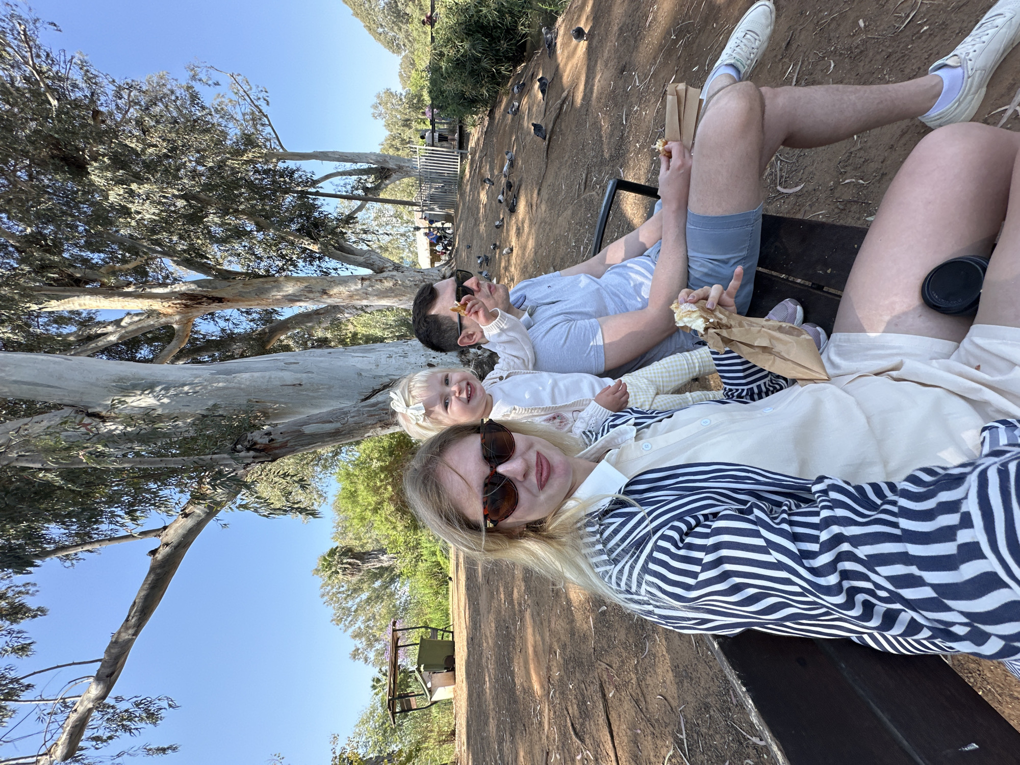 A family of three poses for a photograph on a bench in a Tel-Aviv park. The woman, wearing sunglasses and a striped shirt, is closest to the camera. To her left is a fair-haired child wearing a white sweater and a floral patterned skirt. Behind the child is a man with sunglasses, wearing a light gray t-shirt. The man is holding a bite-sized object in his hand and is eating. All three are seated on a dark wooden bench. A dirt area behind the bench contains a flock of pigeons. In the background are various trees and a light blue sky.