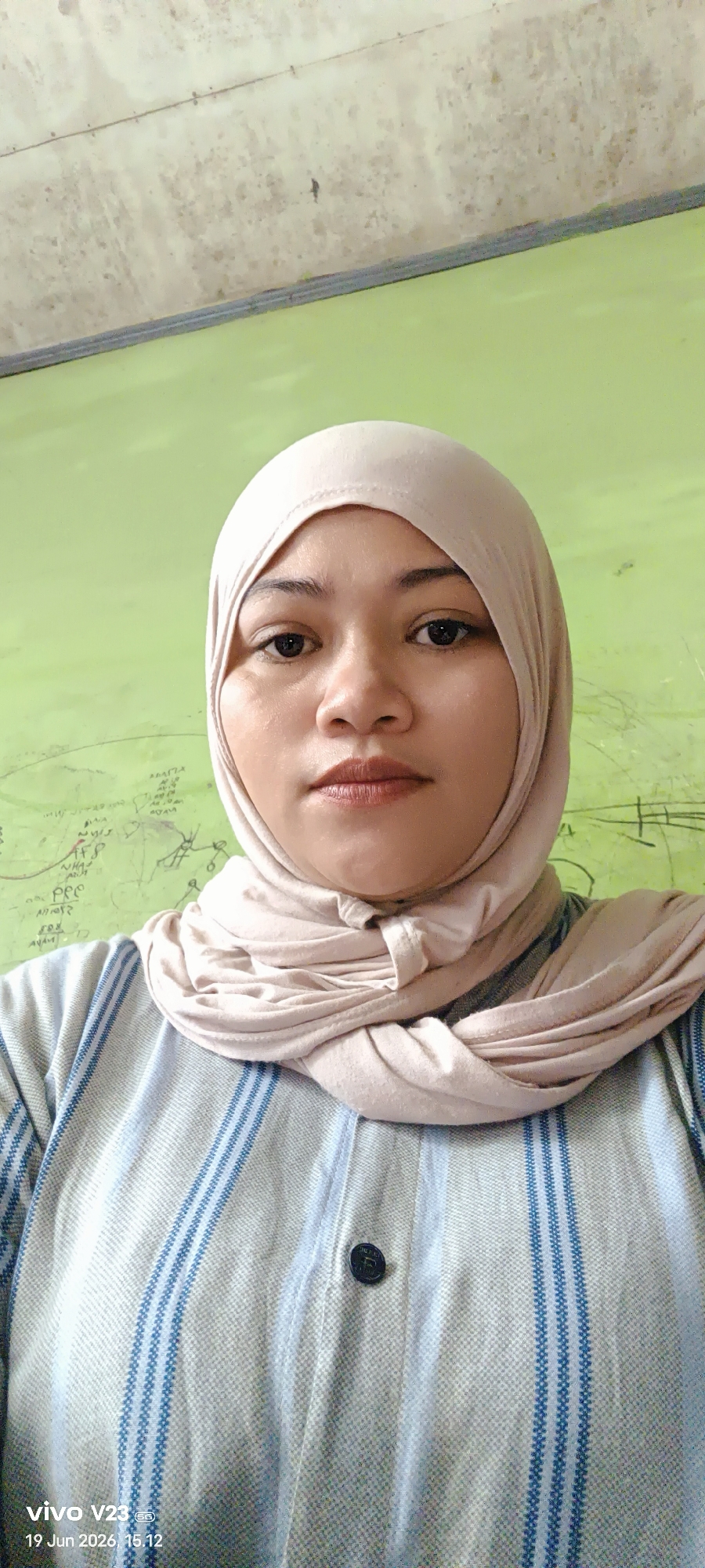 The image shows a medium close-up of a woman wearing a light pink hijab and a striped shirt. She is looking directly at the camera with a neutral expression. The background is a plain, light green wall with some faint markings. A timestamp in the lower left corner indicates "vivo V23, 19 Jun 2026, 15.12". There is no discernible information in the image that places the scene in Pantebayam, Indonesia.