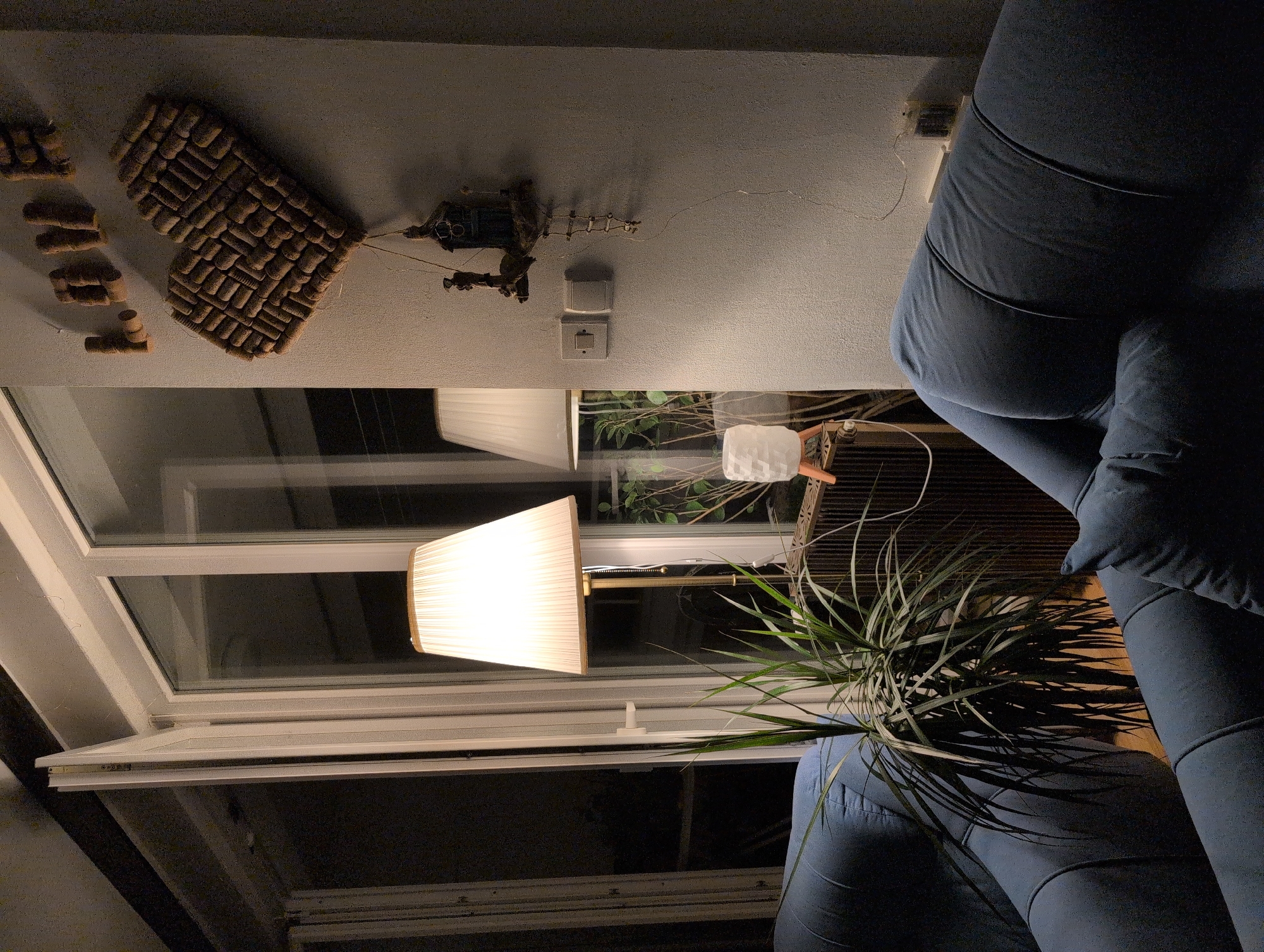The image depicts an interior scene, likely a living room, with a large window and a balcony door partially open. Two blue sofas are visible, one in the foreground and another partially seen on the left. A tall floor lamp with a pleated white lampshade illuminates the space. In the background, a second lamp with a similar shade is visible. A potted plant with long, thin leaves stands between the sofas and the window.
The wall on the right features decorative elements: the word "LOVE" spelled out in what appear to be wine corks, and a heart shape also constructed from wine corks. Below these decorations, a small, rustic-looking decorative item hangs on the wall, resembling a miniature house with a tiny ladder. Two light switches are also present on the wall.
The overall atmosphere is cozy and dimly lit, with warm light from the lamps. The view through the window suggests it is nighttime or dusk, with reflections visible on the glass. The location is identified as Grad Rijeka, Croatia, though no specific landmarks or identifying features of the city are visible within the image itself. The scene is static, with no people or apparent activity.