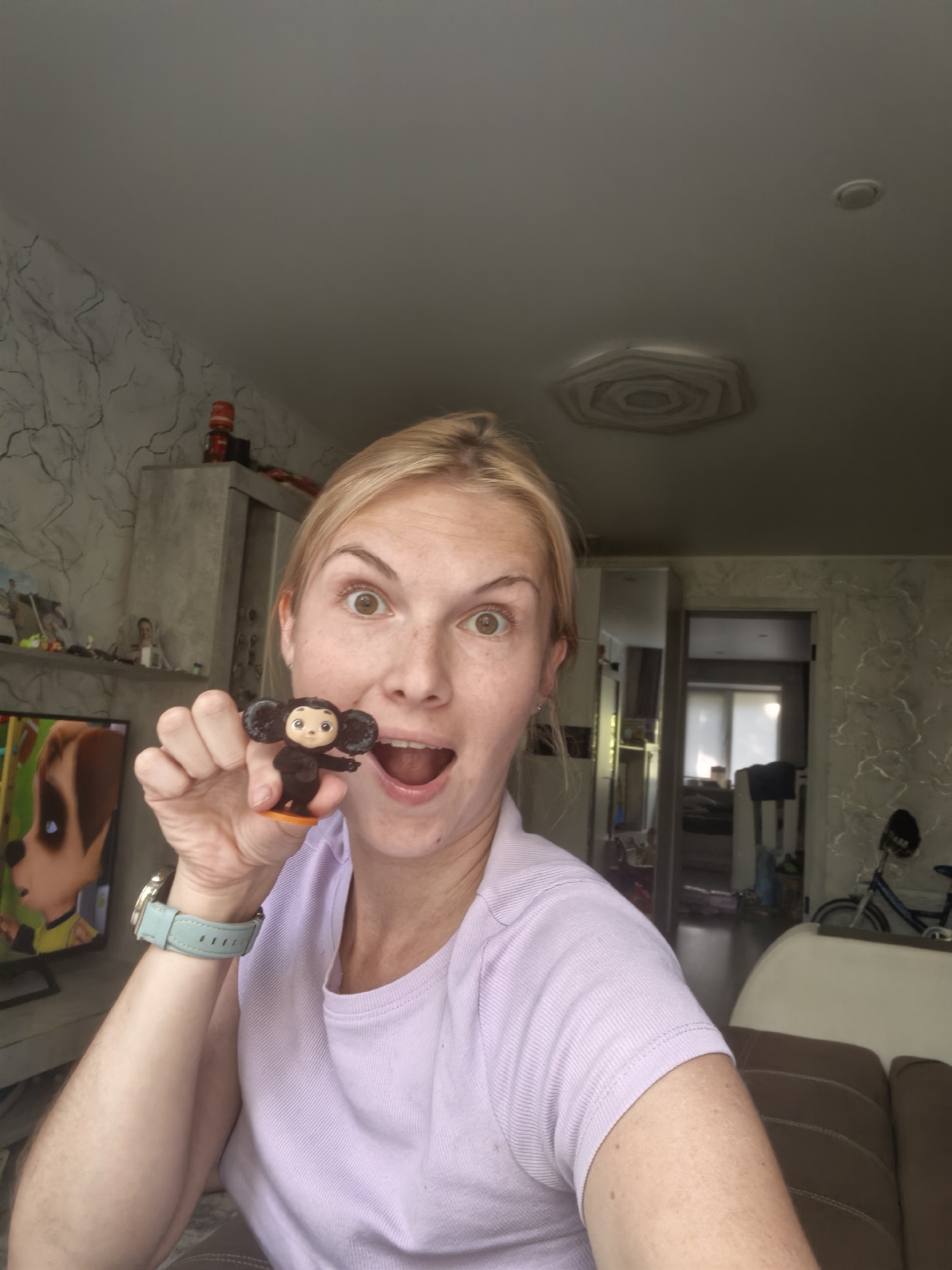 A woman is taking a selfie indoors, holding a small figurine of a character with large ears and dark fur. She is wearing a light purple shirt and a light blue watch on her left wrist. Her mouth is open and her eyes are wide. In the background, there is a television screen showing an animated character that appears to be a dog, a cabinet with items on top, a doorway leading to another room, and a bicycle. The walls have a patterned wallpaper. The ceiling has a textured design in the center and a circular light fixture. The overall impression is a casual home environment.