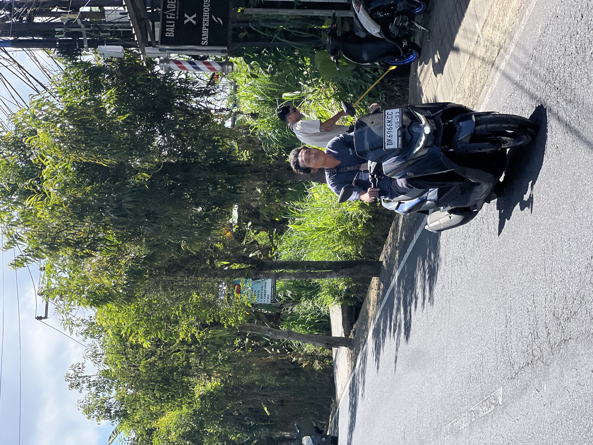A young man rides a dark-colored Yamaha scooter on a paved road in Payangan, Indonesia. He is wearing a dark blue t-shirt with a logo on the left chest. His scooter has a license plate that reads "DK6966KCC". In the background, another man in a light-colored t-shirt is standing near a barber shop called "BALI FADE BARBERSHOP" with a barber pole. Lush green trees and tall grass line the right side of the road, with power lines visible above. The sun is shining, casting shadows on the road.