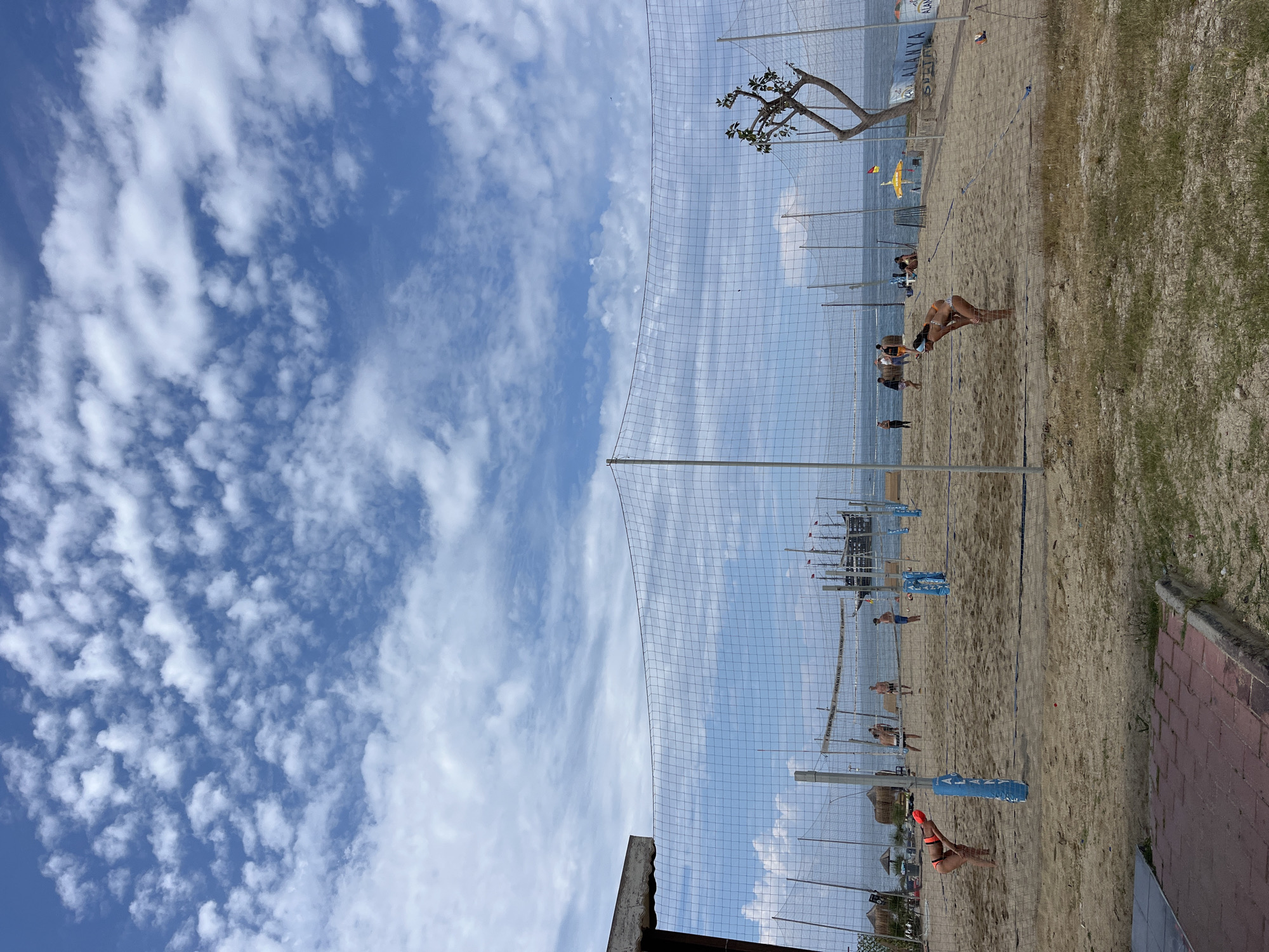The image captures a vibrant scene at Alanya Beach in Turkey. The foreground showcases a beach volleyball court, complete with a net and boundary markers, where people are actively playing. The sky is blue with scattered clouds. The sandy beach transitions into grassy areas, and a brick walkway is visible on the edge. A tree with distinctive branches stands near the court, while the backdrop includes a clear view of the sea. Further down the beach, several pirate ship-themed tourist boats are visible.