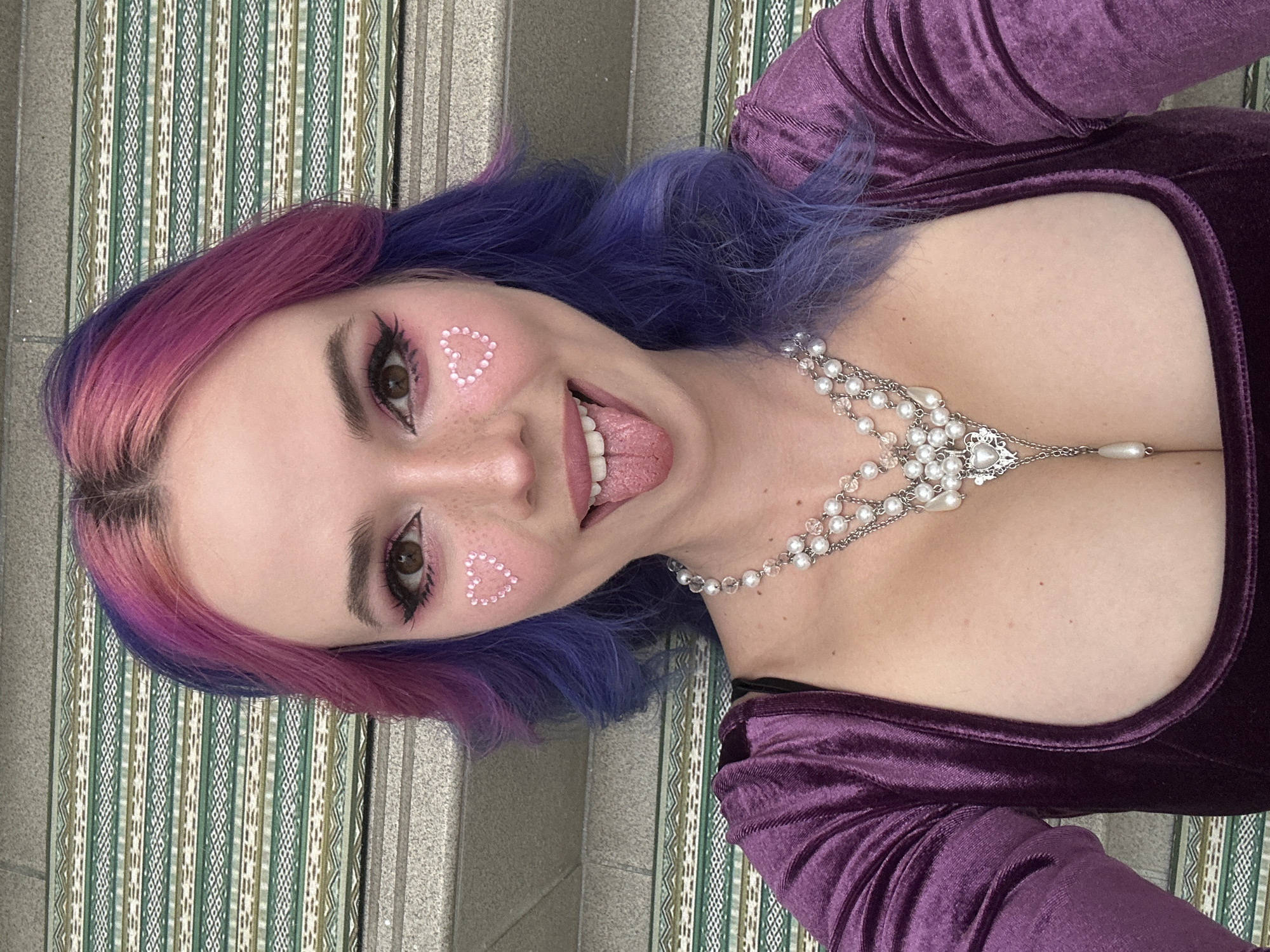 This close-up portrait, captured from a slightly upward angle and presented rotated ninety degrees clockwise, features a young individual with striking two-toned hair that transitions from pink and magenta at the front to a vibrant blue and purple. The person is looking directly at the camera with a playful expression, their tongue slightly extended.
Their face is adorned with expressive makeup, including dramatic black eyeliner, mascara, pink eyeshadow, and delicate heart-shaped embellishments made of small white beads on both cheeks. Freckles are visible across their nose and cheeks. The individual is wearing a deep purple, possibly velvet, long-sleeved top with a low, square neckline. A prominent silver necklace with multiple strands of pearls and clear beads cascades down their chest, featuring an ornate central pendant and a single pearl drop.
The background shows a textured surface, possibly a wall or floor, with vertical striped patterns in shades of green, beige, and white, alongside sections of gray, textured concrete or stone. The overall impression is one of cheerfulness and expressiveness. The context provided indicates this image is from the City of Zagreb, Croatia.