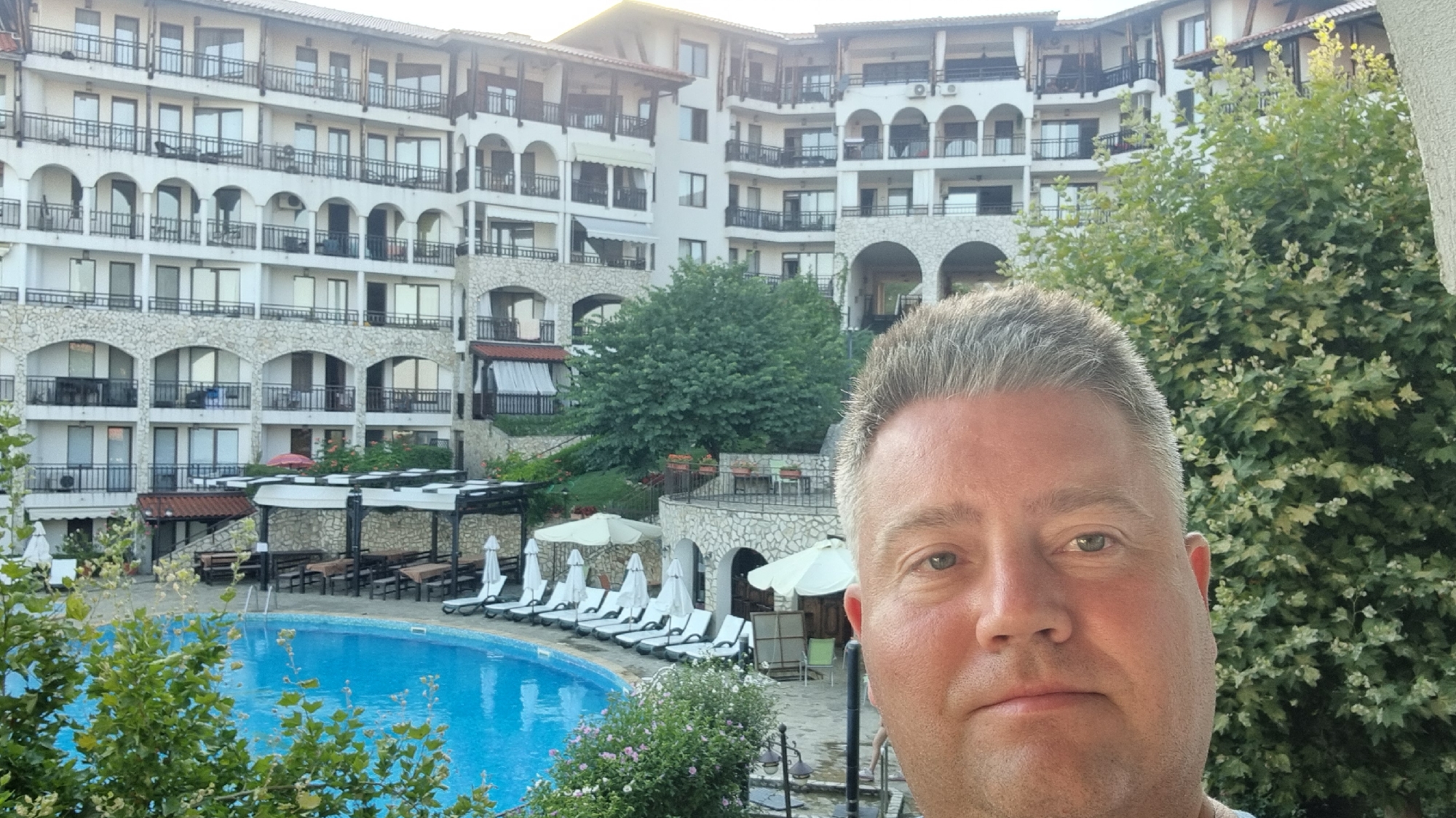 This is a selfie taken by a man in the foreground, with a resort complex and a swimming pool in the background. The resort appears to be a multi-story building with balconies and arches, constructed with white walls and stone accents. In front of the resort, there is a large, bright blue swimming pool with several lounge chairs and umbrellas arranged around it. Some trees and greenery are visible, adding to the outdoor ambiance. The man's face is the primary focus of the foreground. The scene suggests a vacation or leisure setting. The location is identified as Sveti Vlas, Bulgaria.