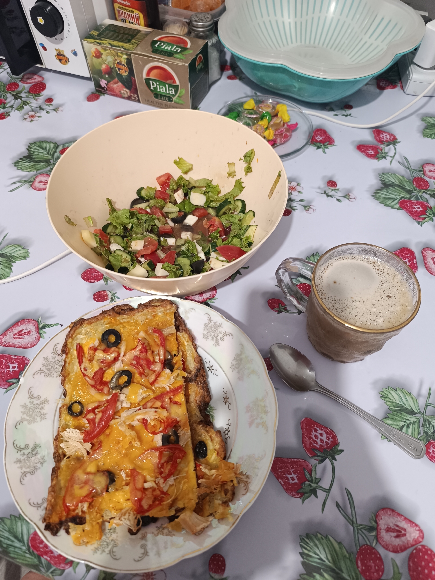 A close-up shot shows a table set for a meal. The table is covered with a tablecloth featuring a strawberry pattern.
In the foreground, a white plate holds a slice of what appears to be an open-faced sandwich or pizza, topped with melted cheese, sliced tomatoes, and black olives. Next to the plate, a spoon rests on the tablecloth.
To the left of the plate, a light-colored bowl contains a salad made with chopped lettuce, tomatoes, cucumbers, and white cheese cubes.
To the right of the salad bowl, a clear glass mug is filled with a frothy beverage, likely coffee or a milkshake.
In the background, there are several objects, including a box of "Piala Lux" tea, a salt shaker, and a colander. There are also some colorful candies in a small glass dish. A white appliance with a dial and buttons is visible in the upper left corner.
The scene appears to be indoors, possibly a kitchen or dining area, with natural light illuminating the table. The overall impression is one of a home-cooked meal or snack.
The visible text includes "Piala Lux" on the tea box, and "BETA TEA" etched on the spoon. The location context states "Taraz city administration, Kazakhstan".