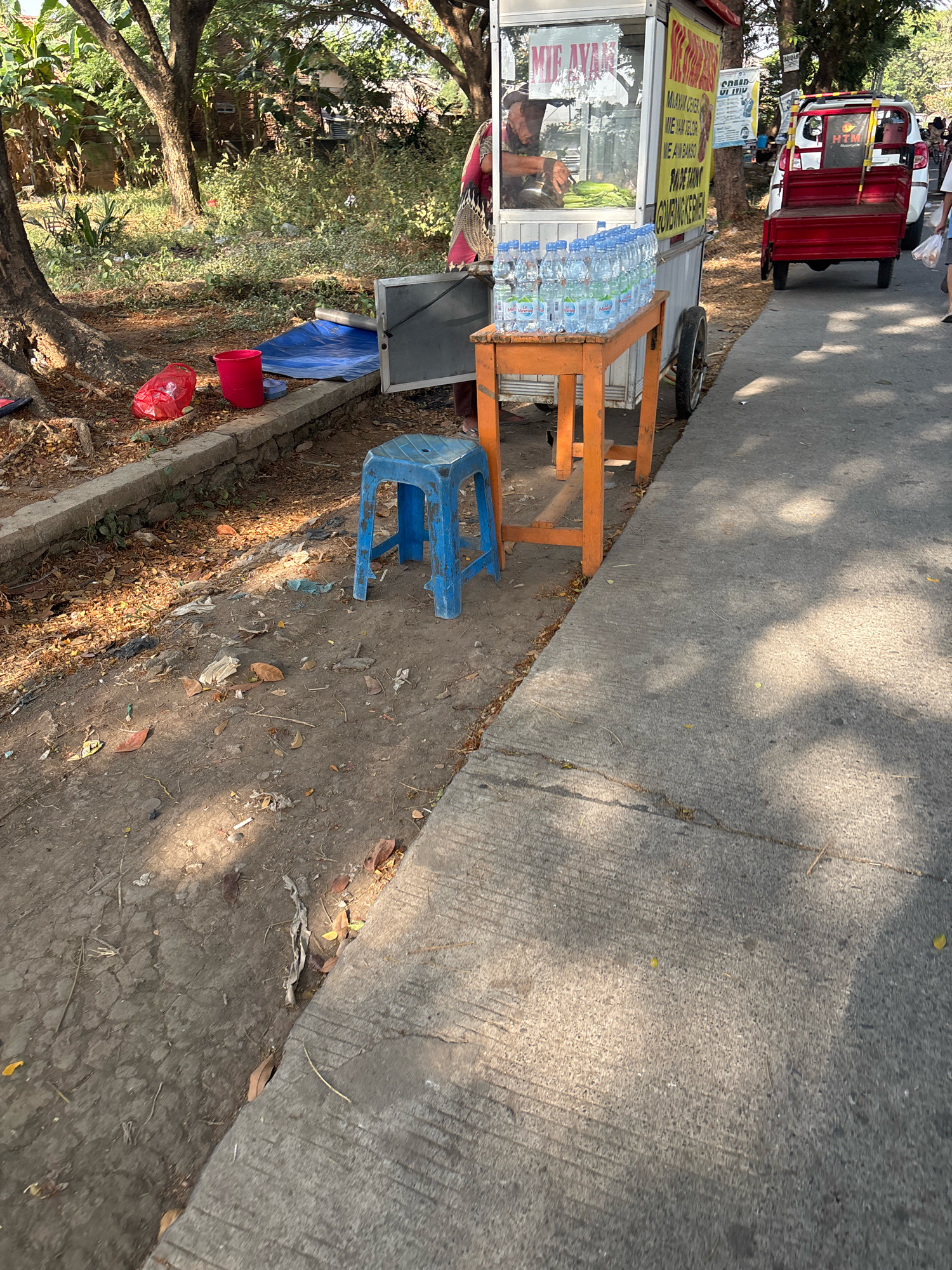 In Telagamurni, Indonesia, on a sunny day, a food cart is parked on the side of a road. The cart, which appears to be selling noodles, has a menu board with text in Indonesian. A person is visible inside the cart, preparing food. In front of the cart, a small orange table is set up with many bottles of water. A blue plastic stool is placed on the dirt and leaf-strewn ground next to the table. A red bucket and a red plastic bag are on the ground near a large tree. A red and white utility vehicle is parked behind the food cart, and other people are walking on the road in the background. The area is surrounded by trees and vegetation, with a concrete sidewalk bordering the road.