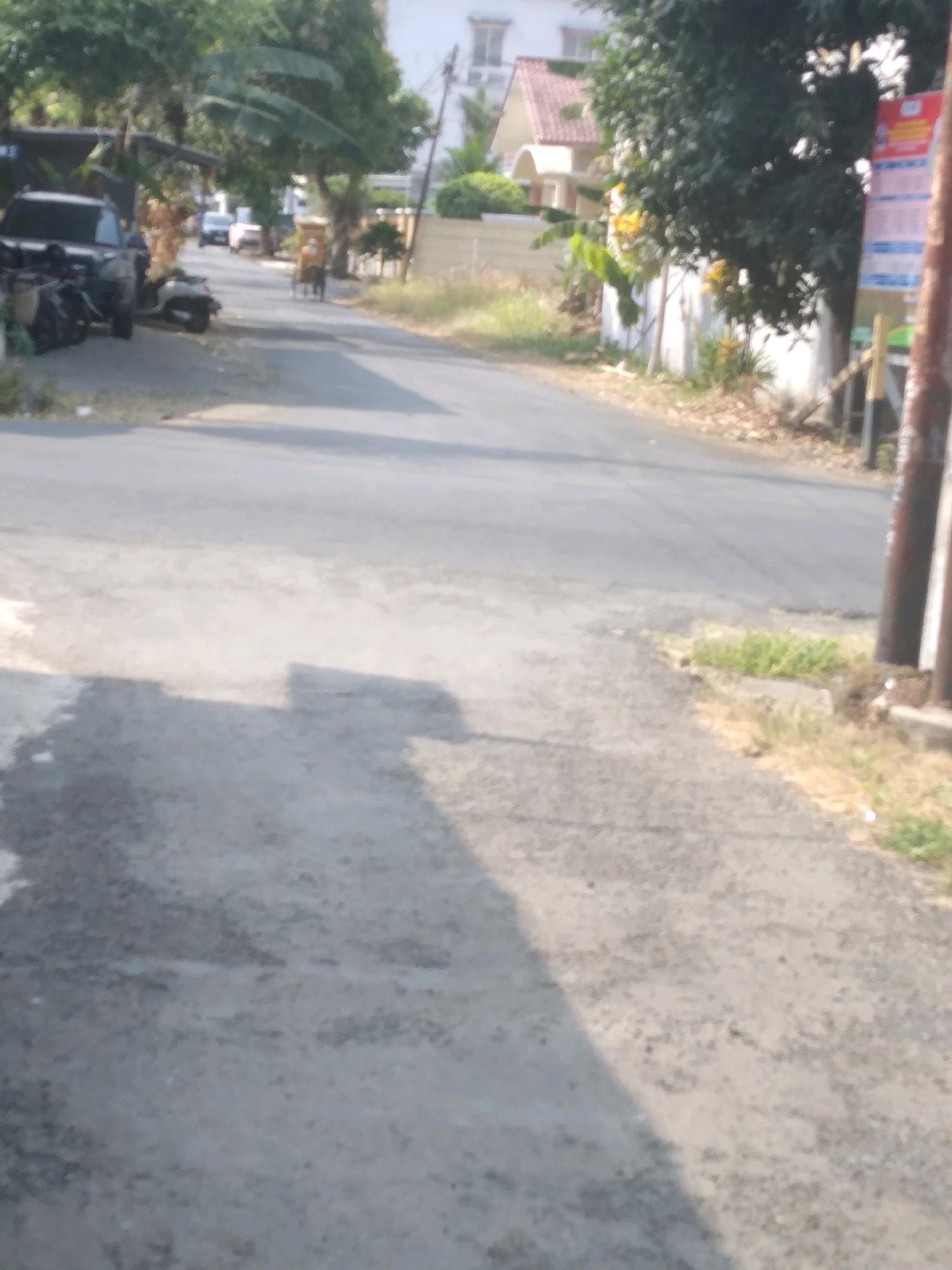 The image depicts a street scene in Desa Pamijen, Kecamatan Sokaraja, Indonesia. A gray asphalt road curves to the right, bordered by dry grass and foliage on the sides. On the left, a dark SUV is parked next to several motorcycles, some of which are covered. In the distance, a person on a bicycle is visible, moving along the road. Trees and houses are present in the background, with one house featuring a red tile roof. A vertical pole with a sign attached is on the right side of the road. The lighting suggests it is daytime, with shadows cast on the road.