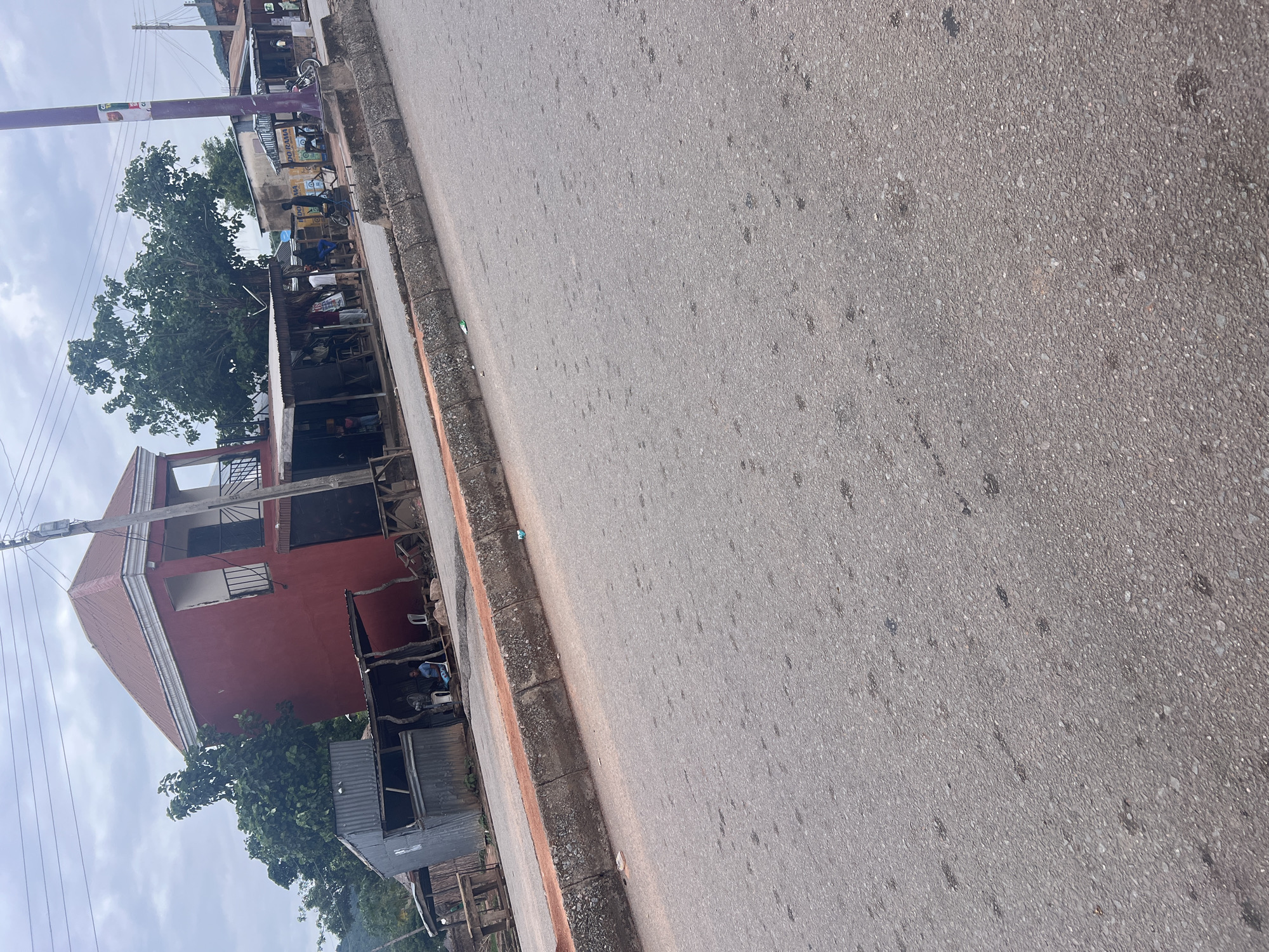 The image captures a street scene in Kona Garu, Nigeria, with a focus on a red building and the adjacent road. The perspective is from the side of the road, looking towards the building and across the asphalt.
**Subjects:**
The scene includes a two-story red building with white trim, featuring balconies with black railings on the upper floor. In front of the building, there are several smaller structures with corrugated metal roofs, some appearing to be stalls or extensions. Trees with dense green foliage are visible behind and around the buildings. The road is paved with asphalt and shows some signs of wear and tear, with scattered dark spots. A few people are visible near the structures, some sitting.
**Activity or Event:**
The presence of people suggests some form of daily activity, possibly commerce or resting. It does not appear to be a major event.
**Notable Details:**
The time of day seems to be daytime, given the lighting. The sky is overcast, indicating cloudy weather. There are power lines visible, suggesting an urban or semi-urban environment. The overall impression is of a somewhat modest and possibly developing area.
**Visible Text:**
Text is visible on a purple pole in the background, but it is too blurry to be legible. There are also some yellow signs on a building in the background with some writing, but it is also not clear enough to read.