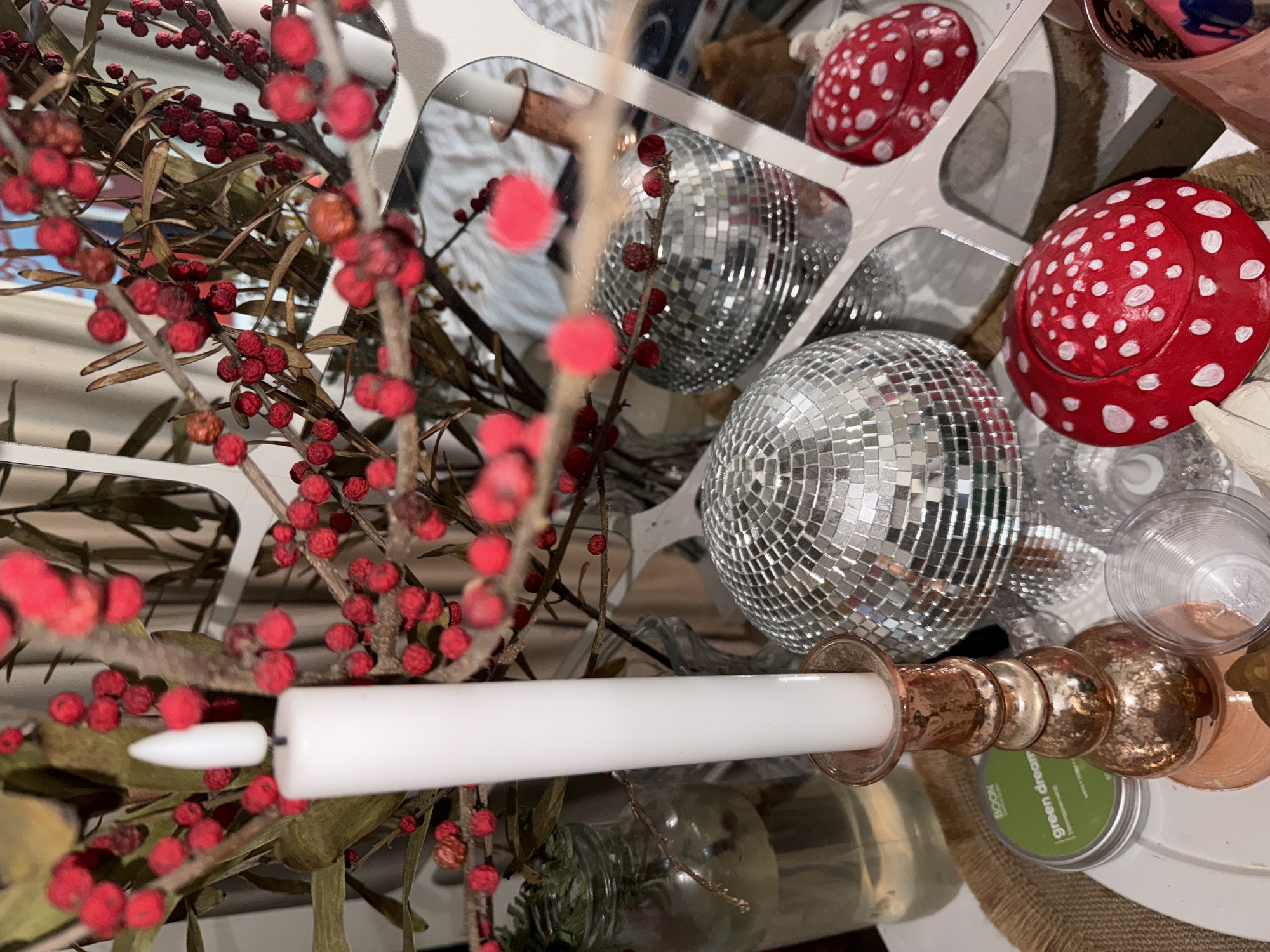 The image presents a still life composition. Predominantly featured are branches with red berries interspersed with elements like disco balls, red-and-white toadstool-shaped decorations, and a white candle in a metallic holder. A partially visible jar and a can with "green dream" text can be seen in the background. The location of this scene is unknown and there is no indication that it is in Krasnoyarsk, Russia.