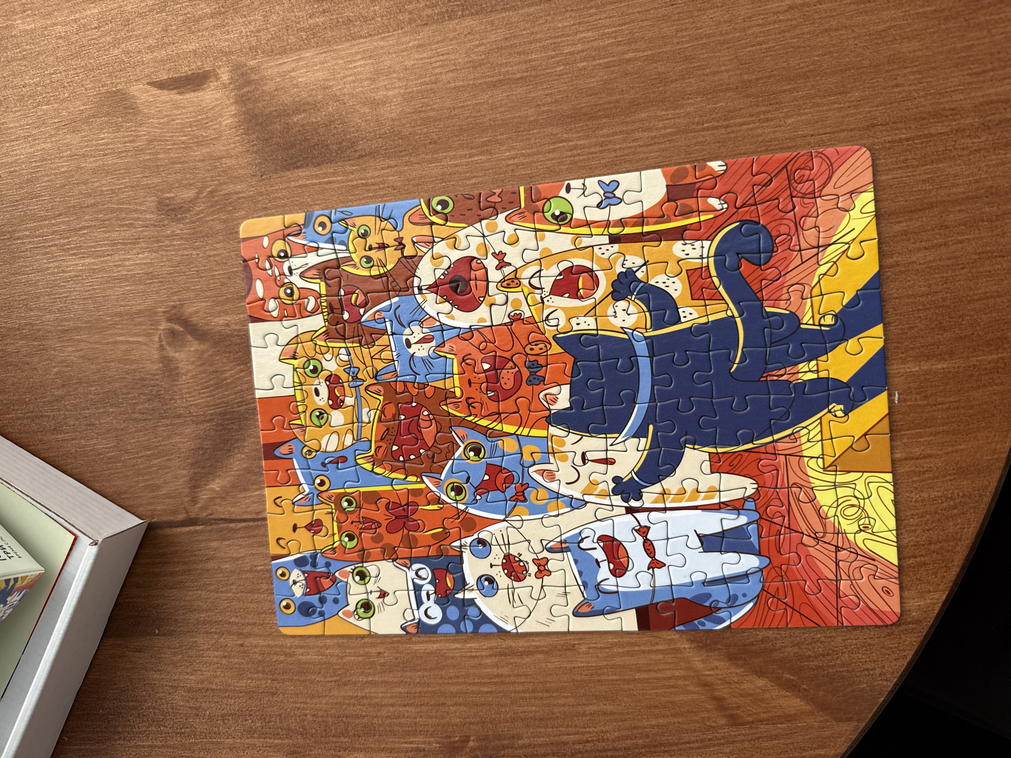 The image depicts a completed jigsaw puzzle on a wooden surface. The puzzle features a colorful, cartoonish illustration of multiple cats stacked on top of each other. The background of the puzzle consists of red, yellow, and orange swirling patterns. A jigsaw puzzle box is partially visible in the lower left corner of the image.
