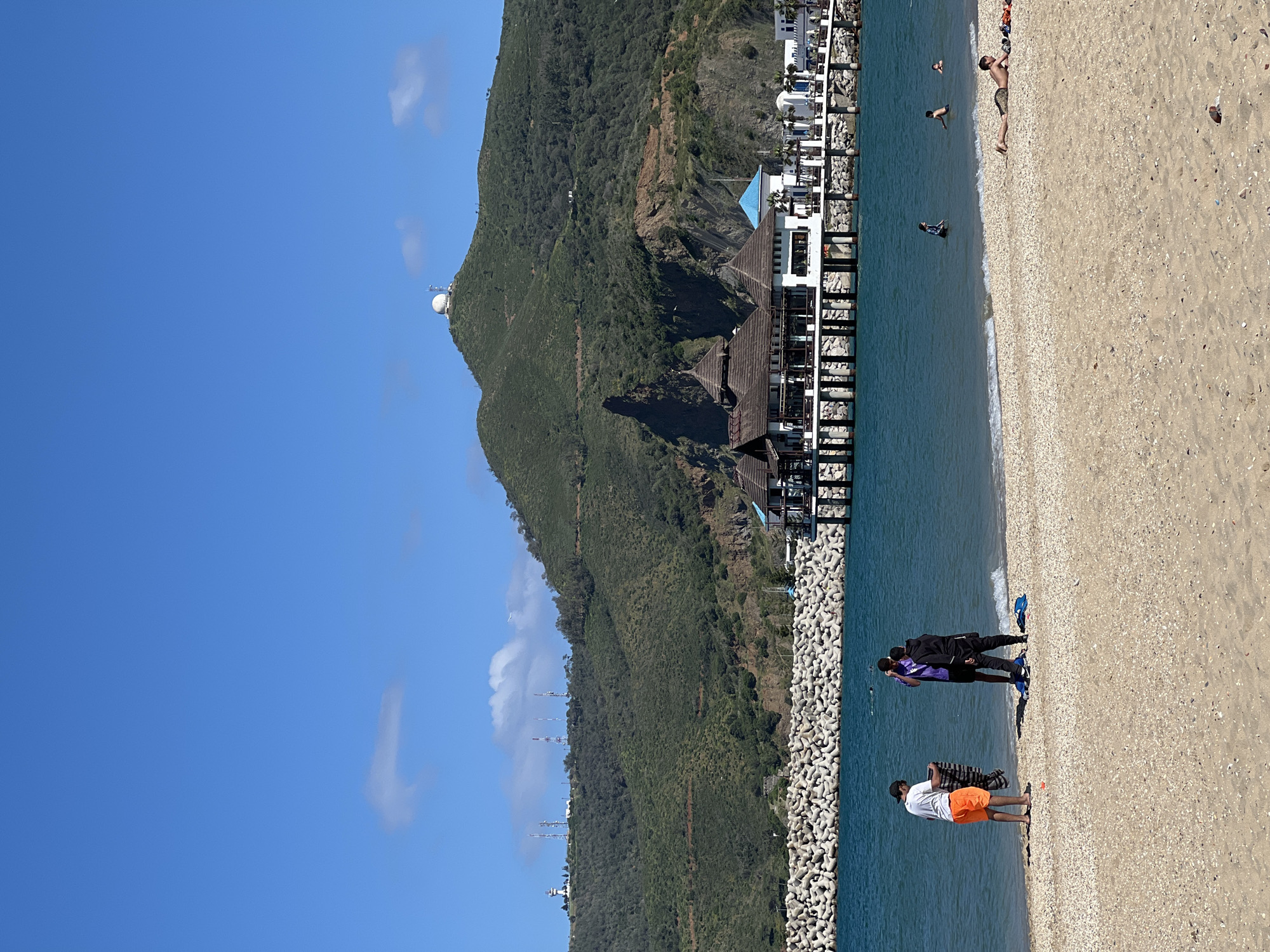 The image depicts a beach scene in M'Diq, Morocco. Clear blue sky occupies the upper left portion of the frame. A tree-covered mountain slopes down to the coast. On the summit is a large, round radar structure. Several communication towers ascend the mountain's flank. At the base of the mountain and extending into the water, a pier structure is visible, partially obscured by dark-brown thatched roofing. White buildings are behind the pier, further up the coast. The shoreline consists of a seawall made of stone tetrapods.
On the sand closer to the viewer, there are several groups of people. One group consists of two figures dressed in dark clothing, with one wearing purple; they are interacting on the shoreline. Nearer to the water, a man wearing a white shirt and orange shorts stands with an item draped over his arm. Further up the beach, other people are visible, including individuals sunbathing. Two people are swimming in the water, a third near the shoreline. The sand is a light tan color with visible shell fragments. The water is a deep blue color. The image has been rotated 90 degrees counter-clockwise.