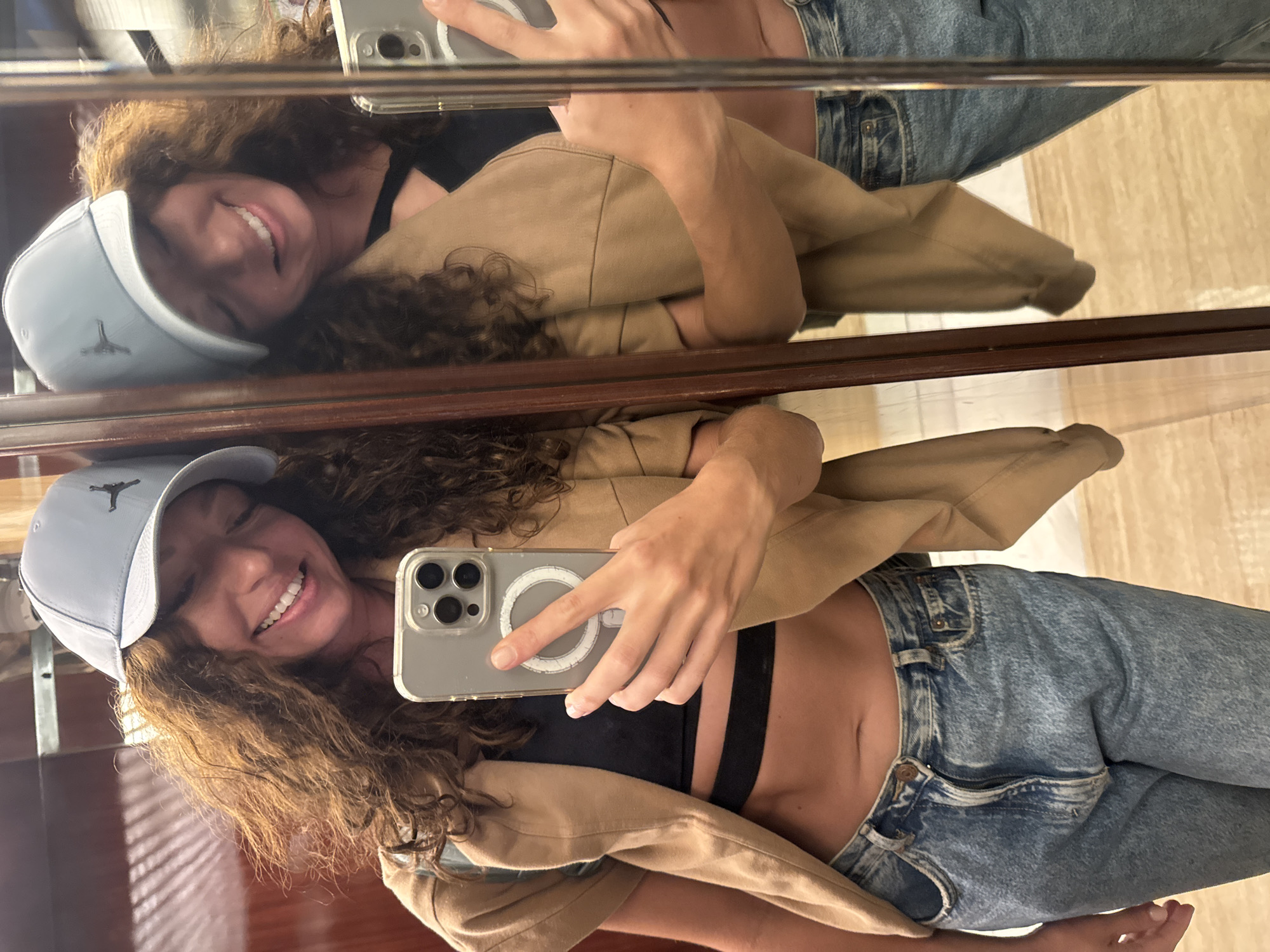 A woman stands indoors, taking a selfie in a mirror. She wears a light blue baseball cap with a black logo, jeans, a black sports bra, and a tan jacket. Her curly hair is brown. She is smiling, holding an iPhone in her right hand. The reflection in the mirror shows a similar image. The background is blurry, suggesting an indoor location. There are no specific identifiers to pinpoint the location in Đà Nẵng, Vietnam. The environment seems clean and modern.
