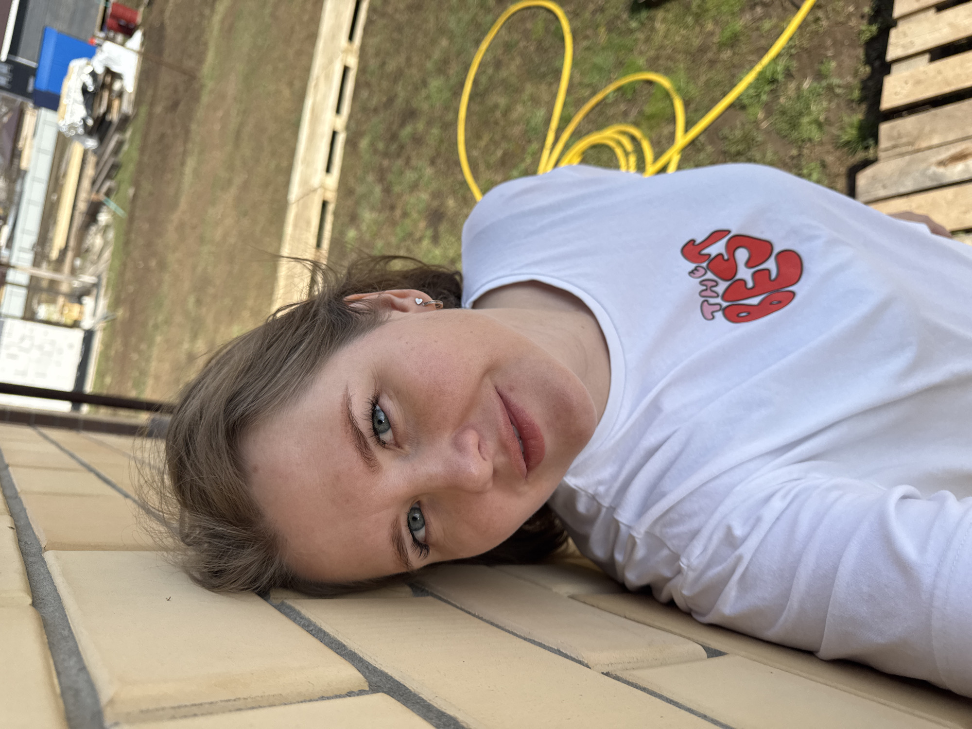 A young woman is depicted in a medium close-up, lying on her side on a light-colored, square-tiled or paved surface, looking directly at the camera. Her head is resting on the surface, and her body extends out of frame to the right, showing her right arm bent at the elbow. She has light brown hair, blue-grey eyes, and a soft, natural smile. She is wearing a plain white long-sleeved t-shirt with a stylized red and pink graphic on the chest, which appears to spell out "THE BEST" or a similar phrase in a bubbly font. She has small earrings, one of which is a heart shape.
The background shows an outdoor setting during what appears to be a bright, sunny day. Beyond the paved surface, there is a grassy area, mostly brown with patches of green, suggesting an early spring or late autumn scene. A coiled yellow garden hose is visible on the grass. Further in the background, to the right, are wooden pallets stacked or arranged, and to the left, blurred building materials, possibly including metal structures and blue tarps, indicate a construction or worksite nearby. The overall impression is one of casual relaxation in an outdoor, semi-developed environment, consistent with a rural or suburban setting in Gornyi (Dvurechenskoe S/P), Russia.