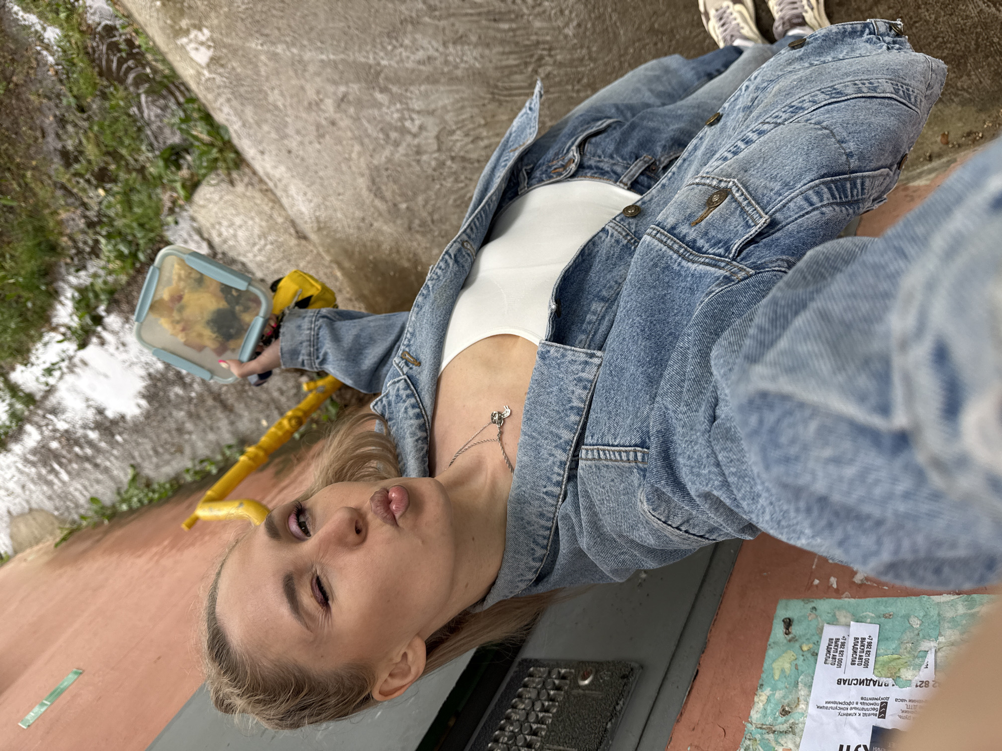Here's a description of the image:
A young, fair-skinned woman is taking a selfie outdoors. She has blonde hair, one eye is partially closed, and she is making a pouting face. She's wearing a white top and a blue denim jacket. A silver necklace is visible on her chest. She's holding a clear plastic food container with some food inside, as well as a yellow object, perhaps a tool or part of equipment.
The setting appears to be an urban area. She's standing near a concrete structure next to some vegetation. On the ground near her are a dark grey object, possibly a metal access panel, and some torn white papers with Russian text. The text includes the phrase "ВЫКУП АВТО" (which means "auto purchase" or "car buyout") and a phone number beginning with "+7 982 821." Given the phone number and the Russian text, it can be inferred that the location is in Russia.