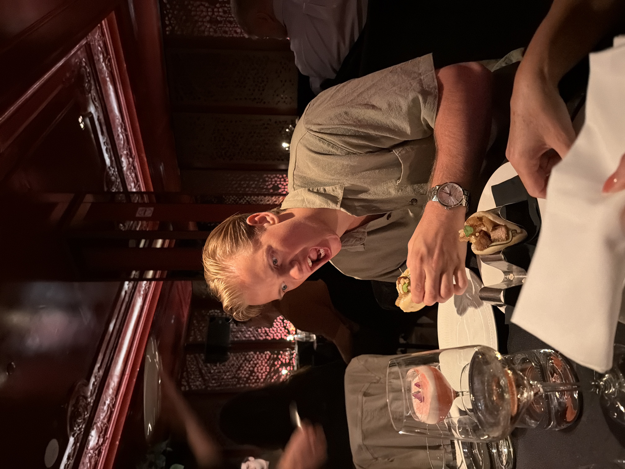 A man with short blonde hair is seated at a table in a dimly lit restaurant. He is wearing a light green button-down shirt and a watch. He appears to be eating or about to eat a small taco or similar appetizer. The table is set with white plates, silverware, and a unique hourglass-shaped cocktail glass filled with a pink and white drink. A white napkin is also on the table. Other people are visible in the background. The restaurant has a dark, ornate wood interior. The location may be in Barcelona, Spain.