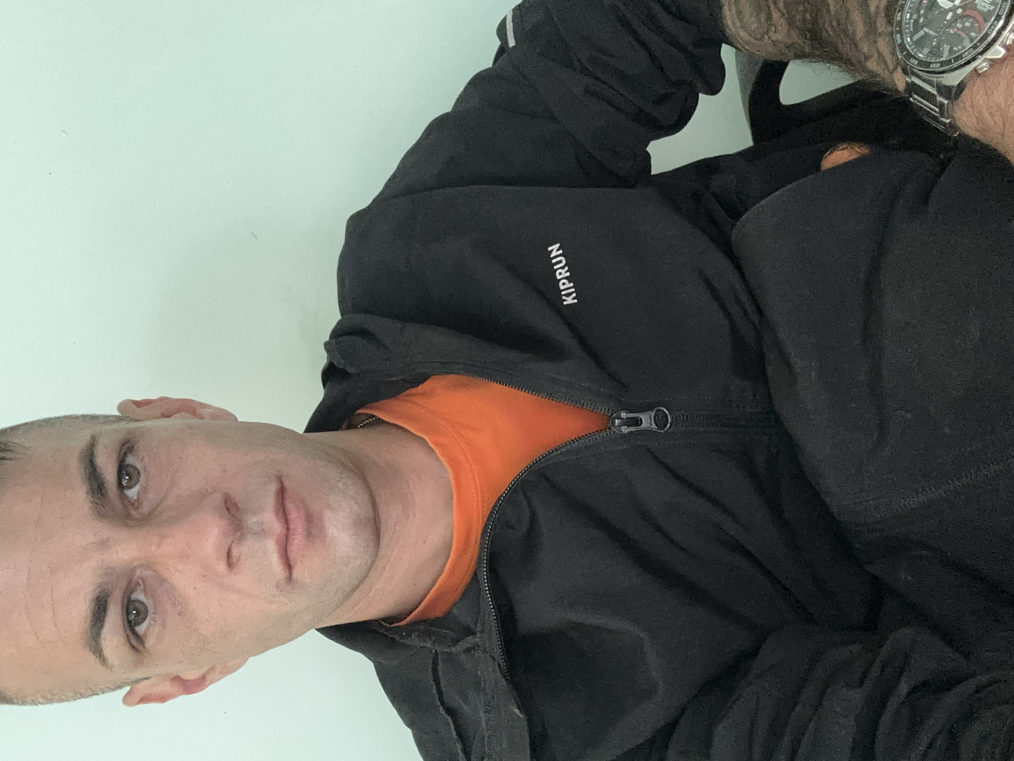 A medium shot photograph captures a fair-skinned man with short, buzzed dark hair and light brown eyes. He is wearing a black zip-up jacket with an orange t-shirt underneath. The word "KIPRUN" is visible on the left chest of the jacket. The man is seated, with his arms crossed. On his left wrist, he wears a silver watch with a black face and multiple dials. The background is a plain, light teal wall. The image appears to be taken indoors. There is no information available that places this image specifically in Piperevo, Bulgaria.