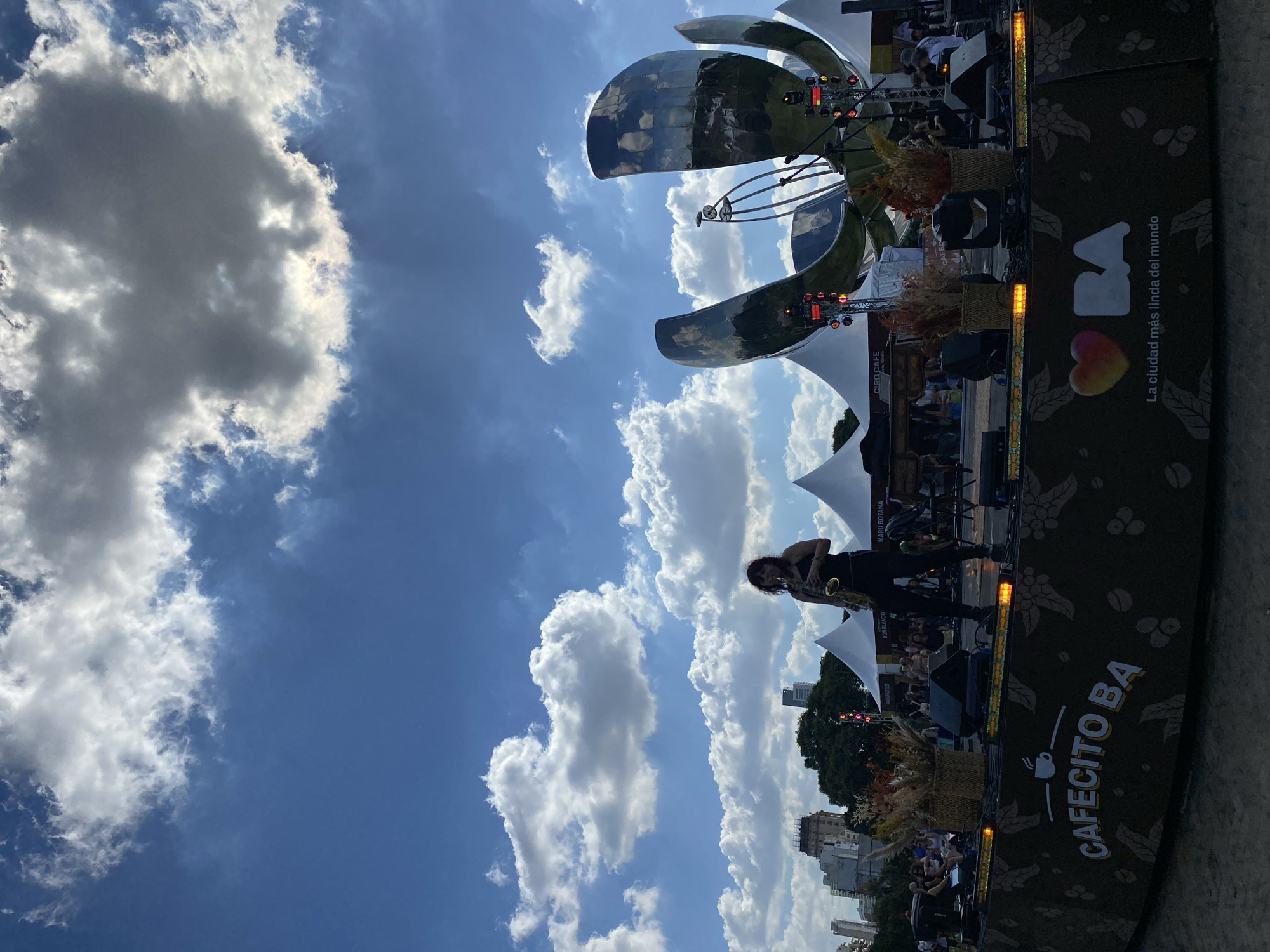 The image captures an outdoor musical performance on a bright, sunny day in Buenos Aires, Argentina. The central subject is a woman playing a saxophone on an elevated stage, partially silhouetted against a clear blue sky adorned with fluffy white clouds.
The stage is set up with professional lighting fixtures, speakers, and decorative baskets filled with dried plants. White tent structures and an audience gathered to watch the performance are visible in the background. Dominating the upper right portion of the scene is the iconic, large metallic flower sculpture, Floralis Genérica, a well-known landmark of Buenos Aires. Beyond the event area, an urban skyline with various buildings can be seen under the bright sky.
The weather is fair, indicative of daytime. The atmosphere appears lively and engaging, consistent with a public cultural event or festival.
Several textual elements are visible. On the stage skirting, the words "CAFECITO BA" and "La ciudad más linda del mundo" (The most beautiful city in the world) are prominently displayed, along with a logo showing "BA" and a heart. Other visible text includes "DON BLANCO," "MARU BOTANA," and "CIRO CAFÉ," likely referring to sponsors or vendors associated with the event. The text "XOXOXX" also appears, though its specific context is unclear.