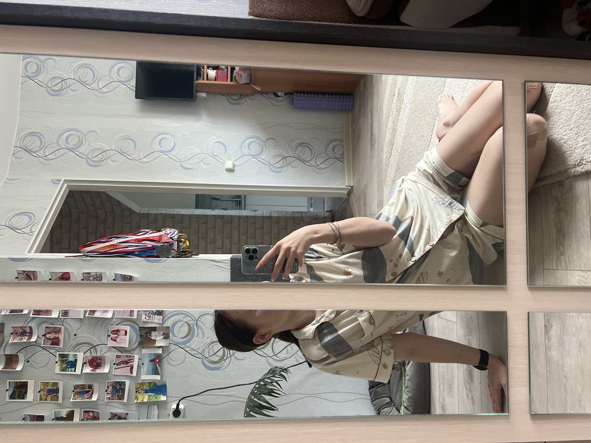 A reflection in a multi-paneled mirror shows a woman in pajamas using a mobile phone. The room has patterned wallpaper, a television, and shelves displaying various items. The woman is wearing a wristwatch. A wall is decorated with pinned photographs. The reflection also shows a partially visible plant. Based on available information, the image is likely taken in Chaikovsky, Russia.