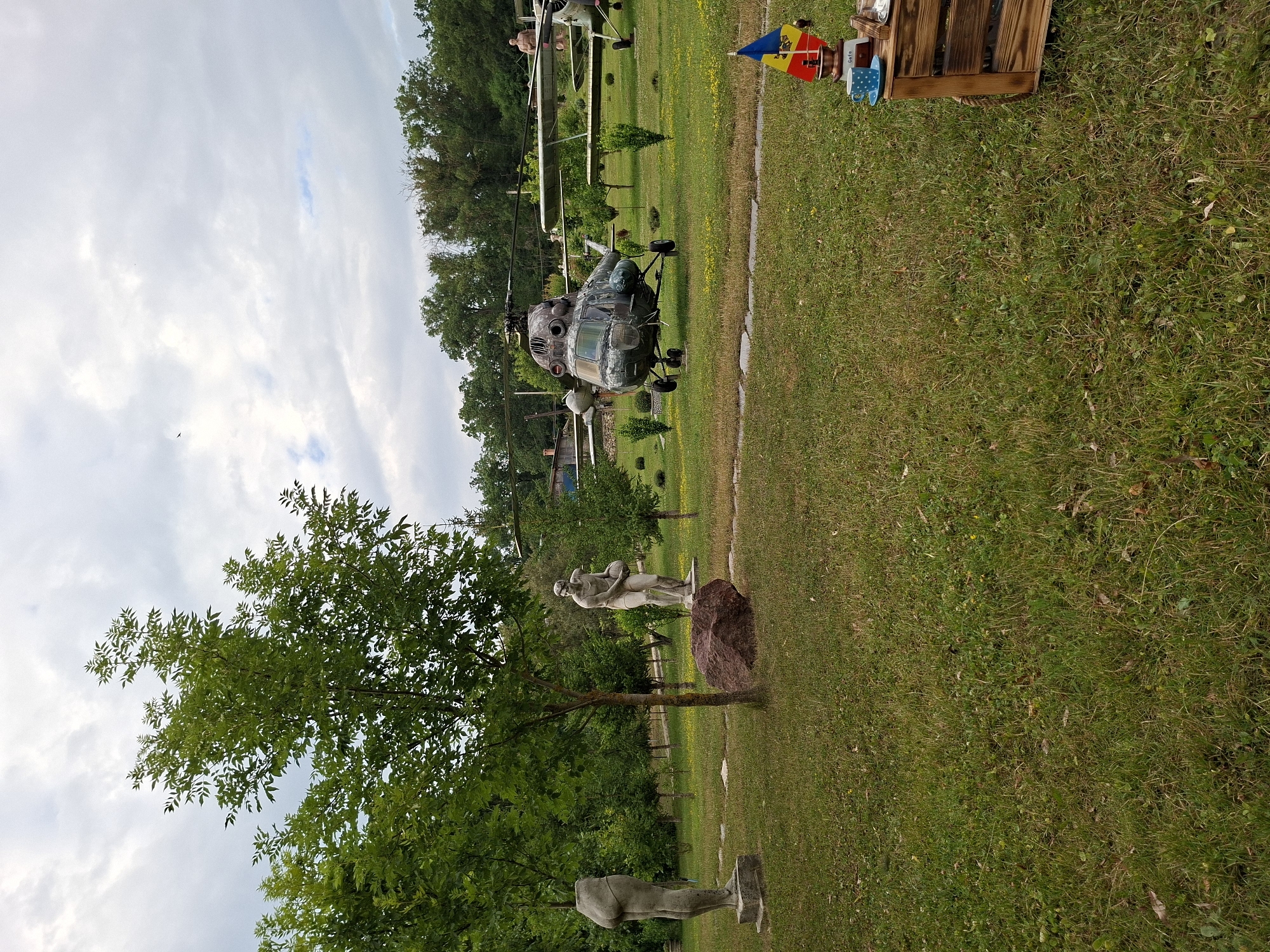 The image depicts an outdoor exhibition space in Malcoci, Moldova, featuring a collection of aircraft and sculptures. In the foreground, a lush green lawn is dotted with scattered leaves. To the right, a rustic wooden crate serves as a display for a small flag of Moldova, a blue coffee cup with white polka dots, a saucer, and a coffee grinder.
Further into the scene, a large, dark-colored helicopter is prominently displayed on the grass. Behind it, a biplane is visible. Several trees, some with dense green foliage, are scattered throughout the area, providing shade and framing the aircraft.
On the left side of the image, two stone sculptures are situated on the lawn. One sculpture depicts the lower half of a human torso, while the other shows a standing male figure holding a ball. A large, reddish-brown boulder rests between the sculptures. In the background, more trees and a distant building are visible under a cloudy sky. The overall impression is of an open-air museum or park dedicated to aviation and art.