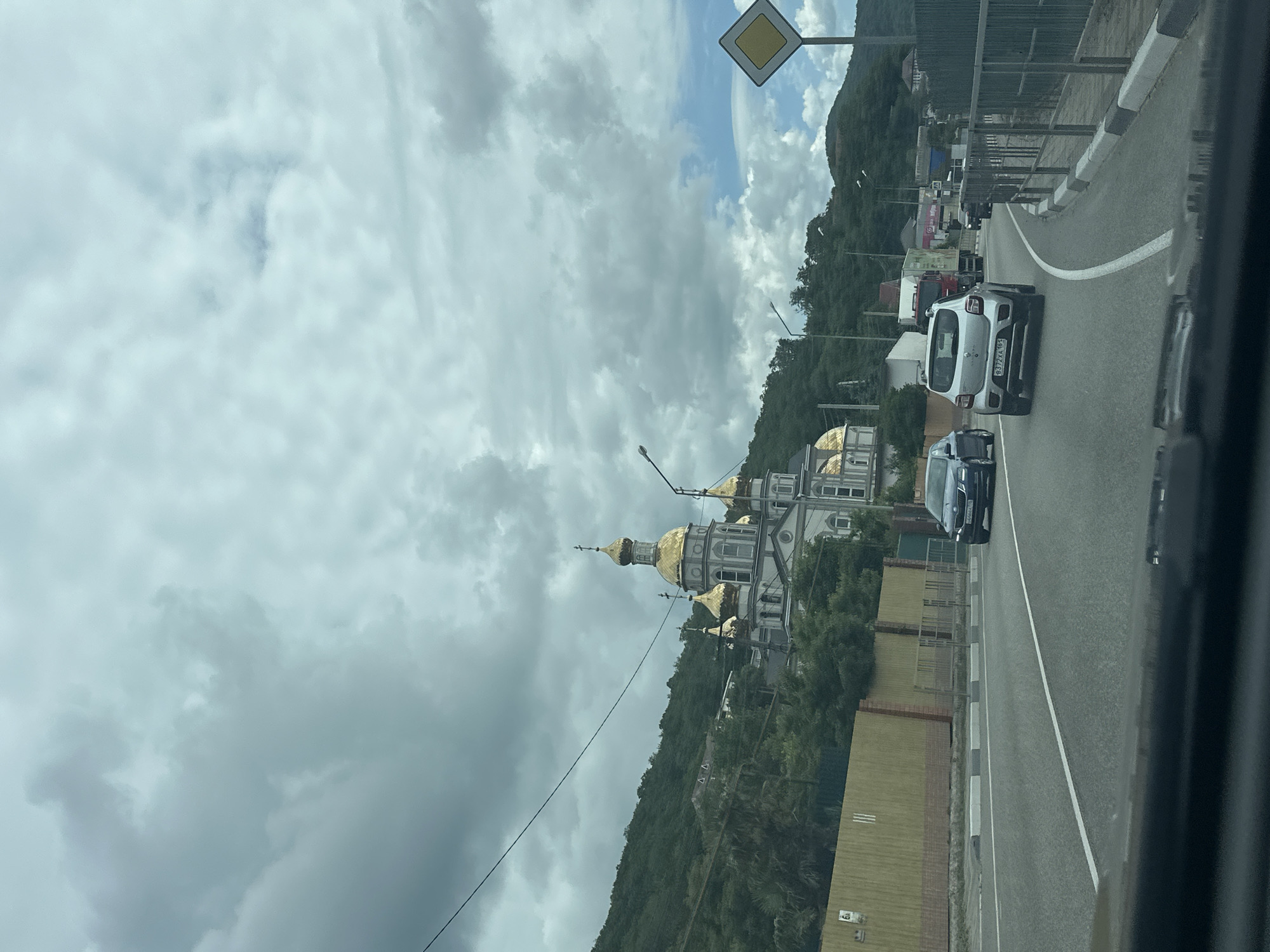 This is a daytime, eye-level, medium shot captured from inside a vehicle. The scene depicts a road with several cars and trucks moving. In the background, a prominent grey and white church with golden domes is situated on a lush green hillside under a cloudy sky. The road is paved with white lane markings and a sidewalk with a metal fence runs alongside it. A yellow diamond-shaped road sign is visible on the right side of the frame. The environment appears to be a coastal town or mountainous region, given the presence of the sea or a large body of water suggested by the hilly terrain. The church is likely a landmark in the area, possibly in or near Olginka, Russia, as suggested by the context.