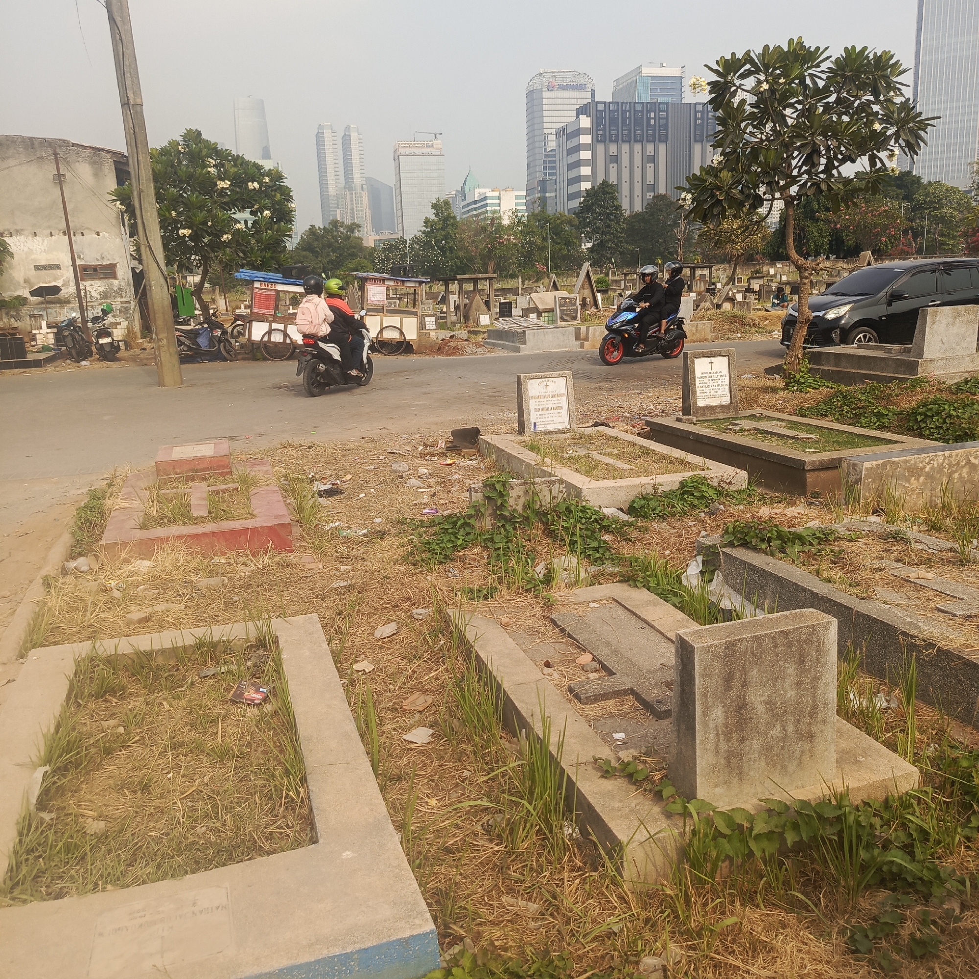 The image depicts a cemetery in South Jakarta, Indonesia, with a backdrop of modern high-rise buildings. In the foreground, several concrete tombstones are visible, some covered with dry grass and weeds. A small tree with white flowers stands to the right of the frame, near the road.
On the road, two people are riding a motorcycle. The rider and passenger are wearing dark clothing and helmets. Further down the road, another motorcycle is approaching, carrying two passengers also in dark attire and helmets. To the left of the road, several parked motorcycles are visible. In the background, a black car is parked. There are also food stalls set up along the road, one with a sign that reads "BAKSO". The overall environment appears to be a blend of urban development and a traditional burial ground, with evidence of dry vegetation and scattered debris around the graves.