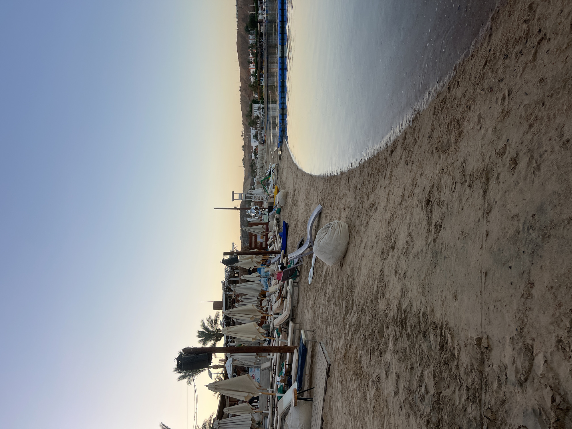 The image depicts a beach scene at sunrise in Sharm El Sheikh, Egypt. The sky is clear with a gradient of soft blue and pale yellow, indicating the early morning light. The calm, shallow water of the sea meets the sandy shore, with a blue floating barrier delineating a section of the water.
The beach is lined with rows of empty lounge chairs and closed umbrellas, suggesting that the day has not yet begun or is drawing to a close. A large, white beanbag chair and a rolled-up white mat are placed on the sand near the water's edge. In the background, a line of buildings and palm trees can be seen against a gently sloping hill, typical of the coastal landscape in Sharm El Sheikh. The overall atmosphere is tranquil and serene, emphasizing the peaceful start or end of a day at the beach. There is no visible text in the image.