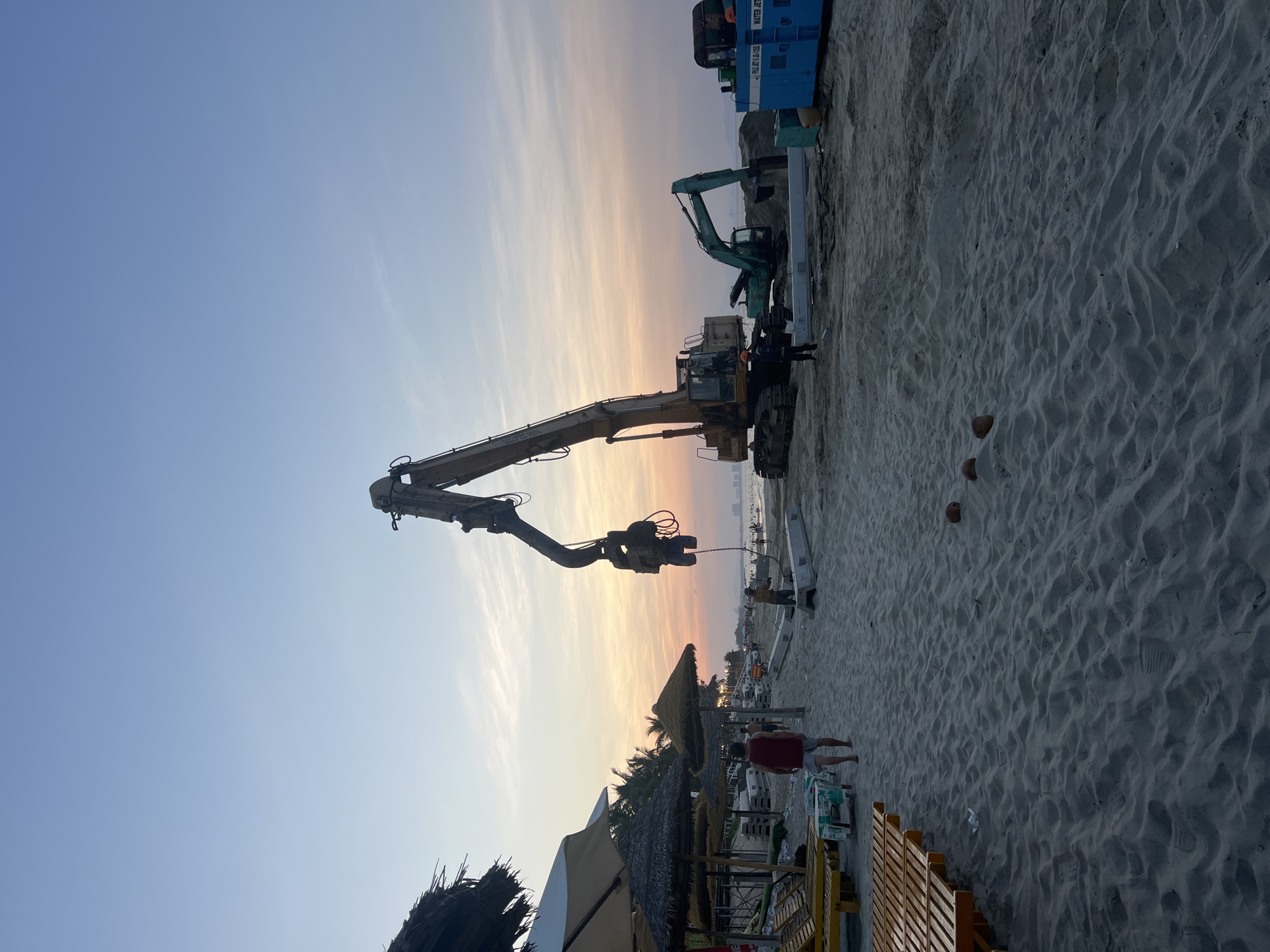 The image depicts a construction scene on a sandy beach in Hoi An, Vietnam, at dusk. A large yellow excavator with a long boom is positioned prominently on the sand, with a smaller green excavator situated behind it. Several white concrete barriers and pipes are laid out on the beach. A blue generator unit is visible to the right.
In the foreground, the sand is marked with footprints and a few coconuts. To the left, several thatched umbrellas and lounge chairs suggest a resort or seating area for beachgoers. A few people are visible walking on the beach, including one person in a red shirt and shorts. The sky displays a gradient of pale blue and orange hues, indicative of sunset. In the distance, the coastline with buildings and a body of water can be seen. The overall scene suggests ongoing construction work on a beach in Hoi An.