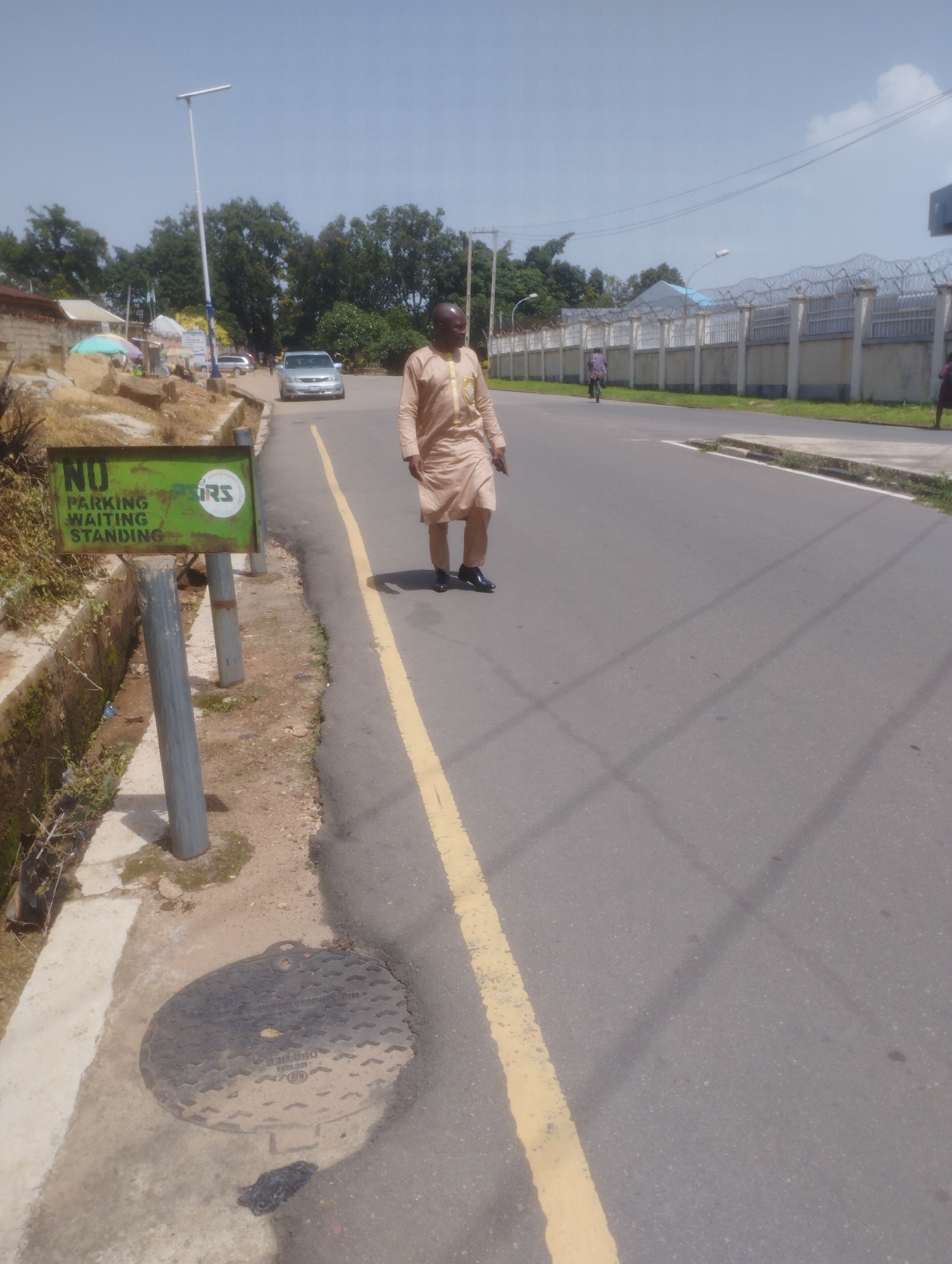 A man in a light brown robe is walking on a paved road in Jos, Nigeria, towards the right of the frame. He is wearing black shoes. The road has a yellow line marking its edge. On the left side of the road, there is a green sign that reads "NO PARKING WAITING STANDING". Next to the sign is a manhole cover. In the background, there are trees, a street light, a building with a fence topped with barbed wire, and several cars parked on the side of the road. The sky is mostly clear with some clouds.