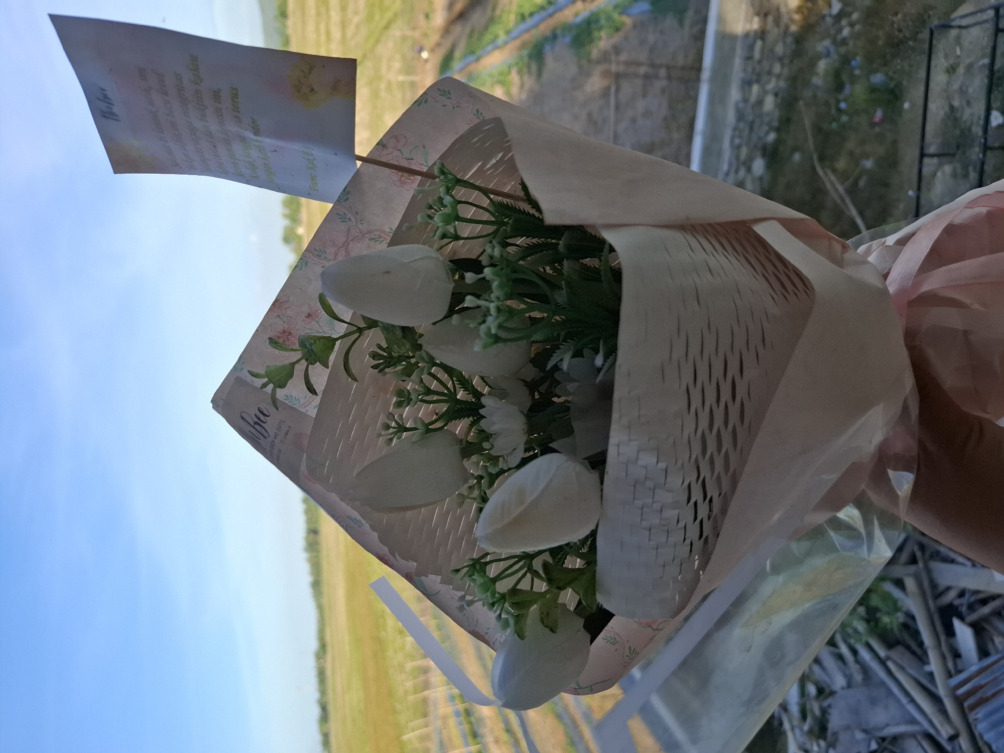 An indoor close-up shot shows a bouquet of artificial white tulips with green foliage. The bouquet is wrapped in patterned paper and a light pink paper cone, tied with a pink ribbon. A small card with handwritten text in Indonesian is inserted into the bouquet. The background reveals a blurred view of a rural landscape with green fields and a blue sky. The image is taken from inside, possibly through a window, with a glimpse of some debris in the lower left corner. The scene is likely set in Gronggrong, Indonesia, given the presence of Indonesian text on the card and the rural scenery.