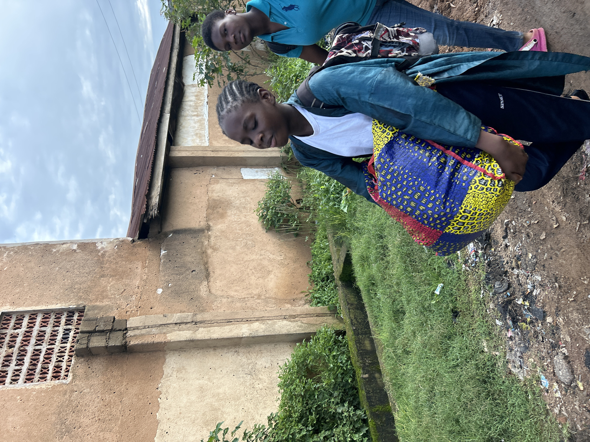 This is a full-shot, eye-level, vertical, outdoor, natural light photograph taken in Jos, Nigeria. The image features two young Black females walking. The female in the foreground is closer to the camera and is facing towards the right. She has braided dark hair and is wearing a white t-shirt under a teal-colored robe, black pants with white stripes, and a backpack with a colorful pattern. She is carrying a large, brightly colored bag in her arms. The second female is behind her, to the right, and is also facing towards the right. She has short dark hair and is wearing a blue polo shirt, blue jeans, and pink sandals. She is also wearing a backpack.
The environment appears to be a city street with buildings and vegetation. To the left of the females, there is a tan-colored building with a patterned window. The ground in front of the building is covered with green grass, a concrete channel, and then a dirt path with some debris. The background shows more buildings and trees, with a cloudy sky overhead. The overall scene depicts a moment of daily life in Jos, Nigeria, with the two girls likely on their way to or from school or another activity.