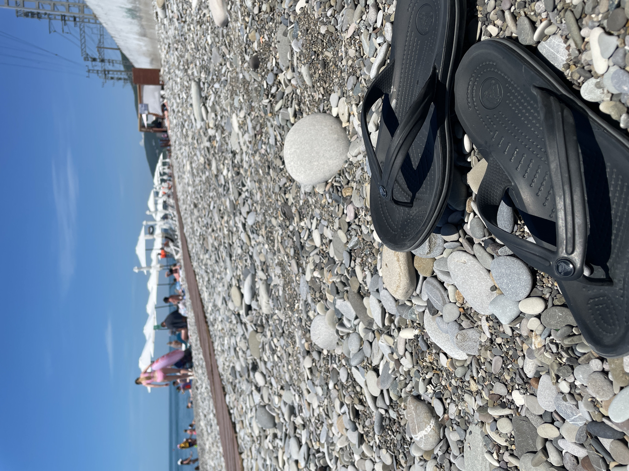A close-up, low-angle shot captures two black flip-flops resting on a pebbled beach. The flip-flops are positioned in the foreground, with one lying slightly behind and to the right of the other. The beach is covered in a variety of smooth, rounded pebbles in shades of grey, white, and brown. In the background, the ocean is visible under a clear blue sky. Further in the distance, a line of white beach umbrellas and several people are present, suggesting a public beach setting. Some infrastructure, including power lines and a concrete wall, are visible on the far right of the frame. The overall scene is sunny and appears to be a typical summer day at the beach.